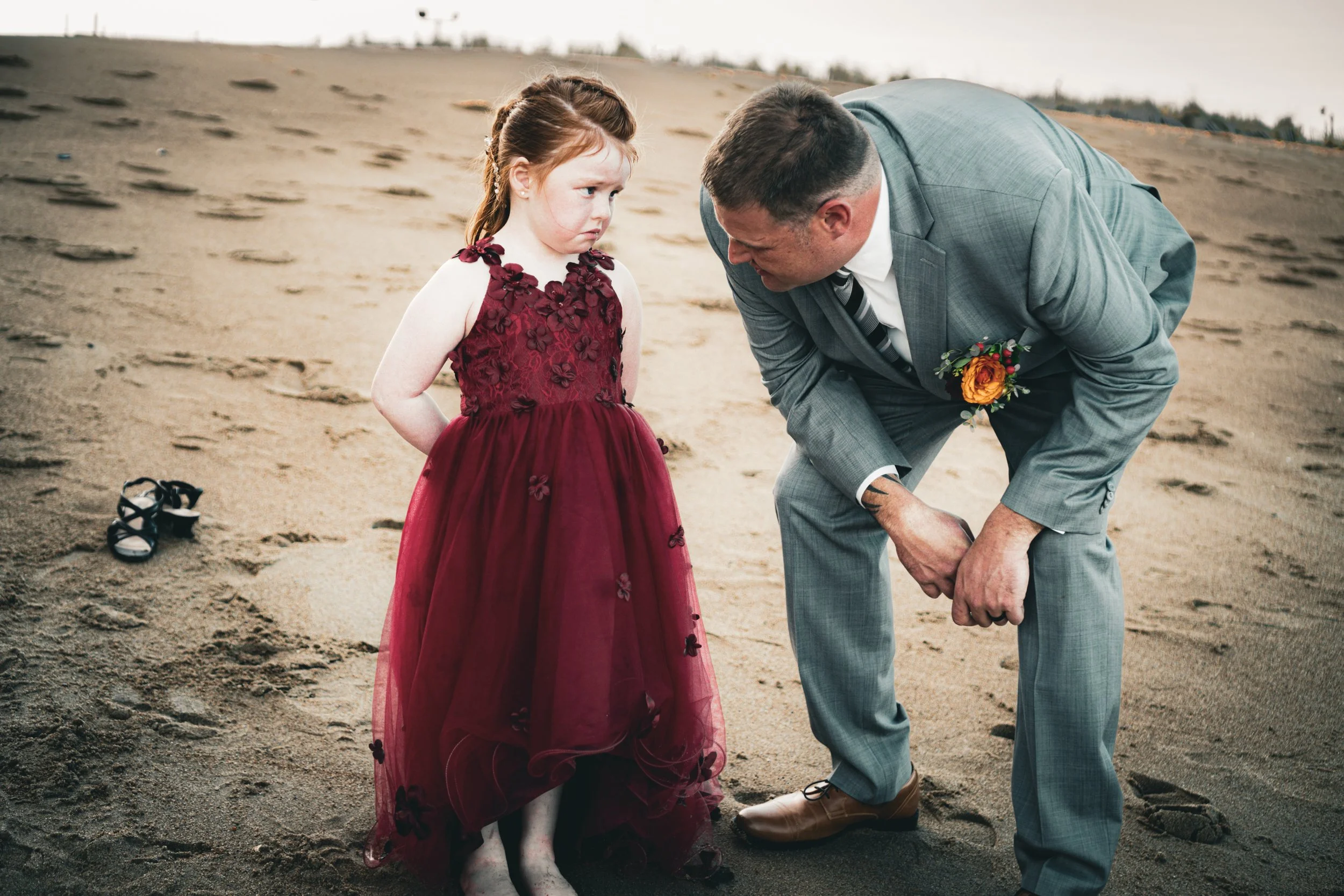 A man in a gray suit with a boutonniere is kneeling on a sandy beach in front of a young girl in a burgundy dress, appearing to tie her shoe or address her.