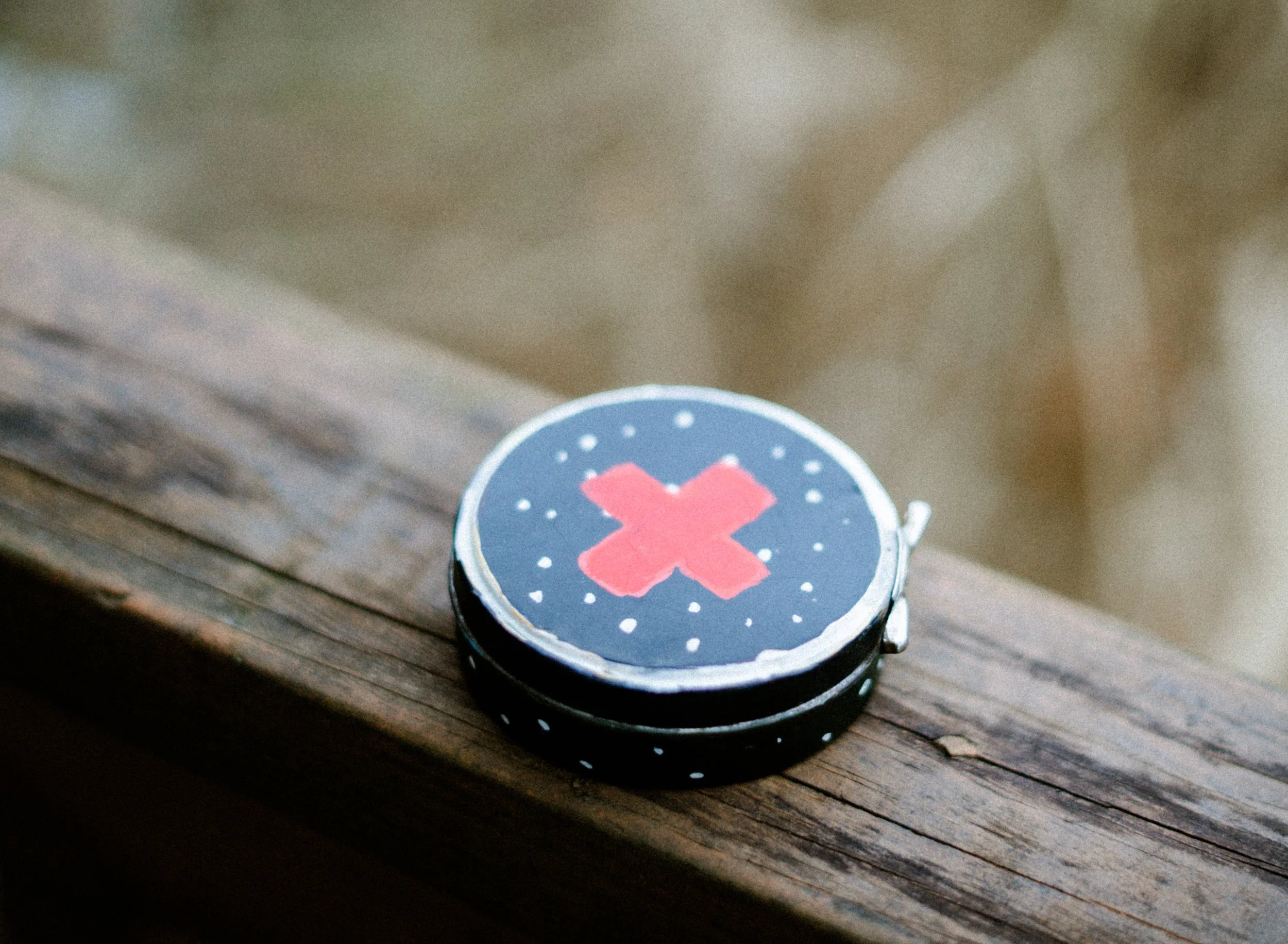 A small round container with a black lid featuring a pink X and white dots, placed on a weathered wooden surface.