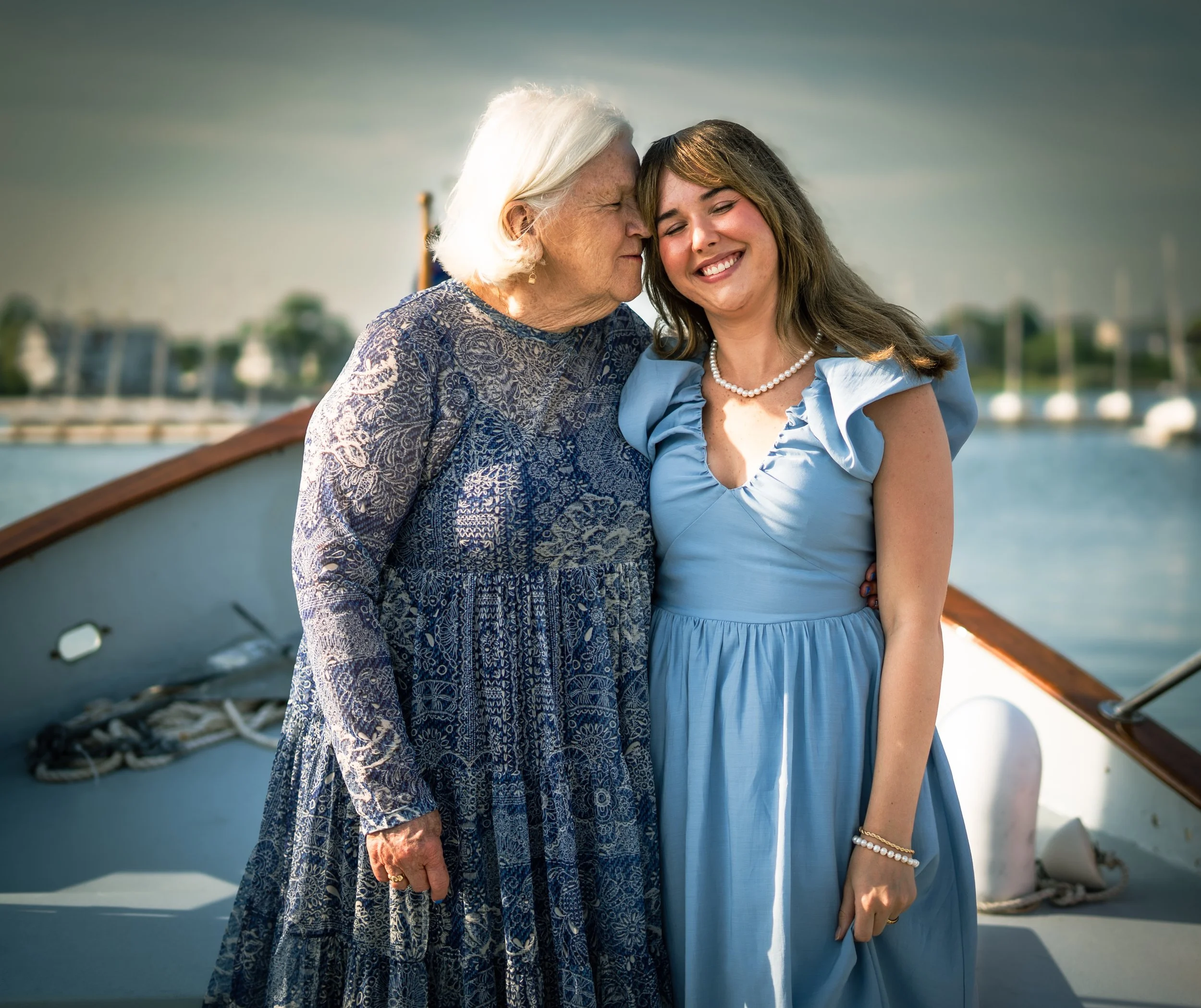 An elderly woman and a young woman standing on a boat by the water, smiling and touching foreheads.