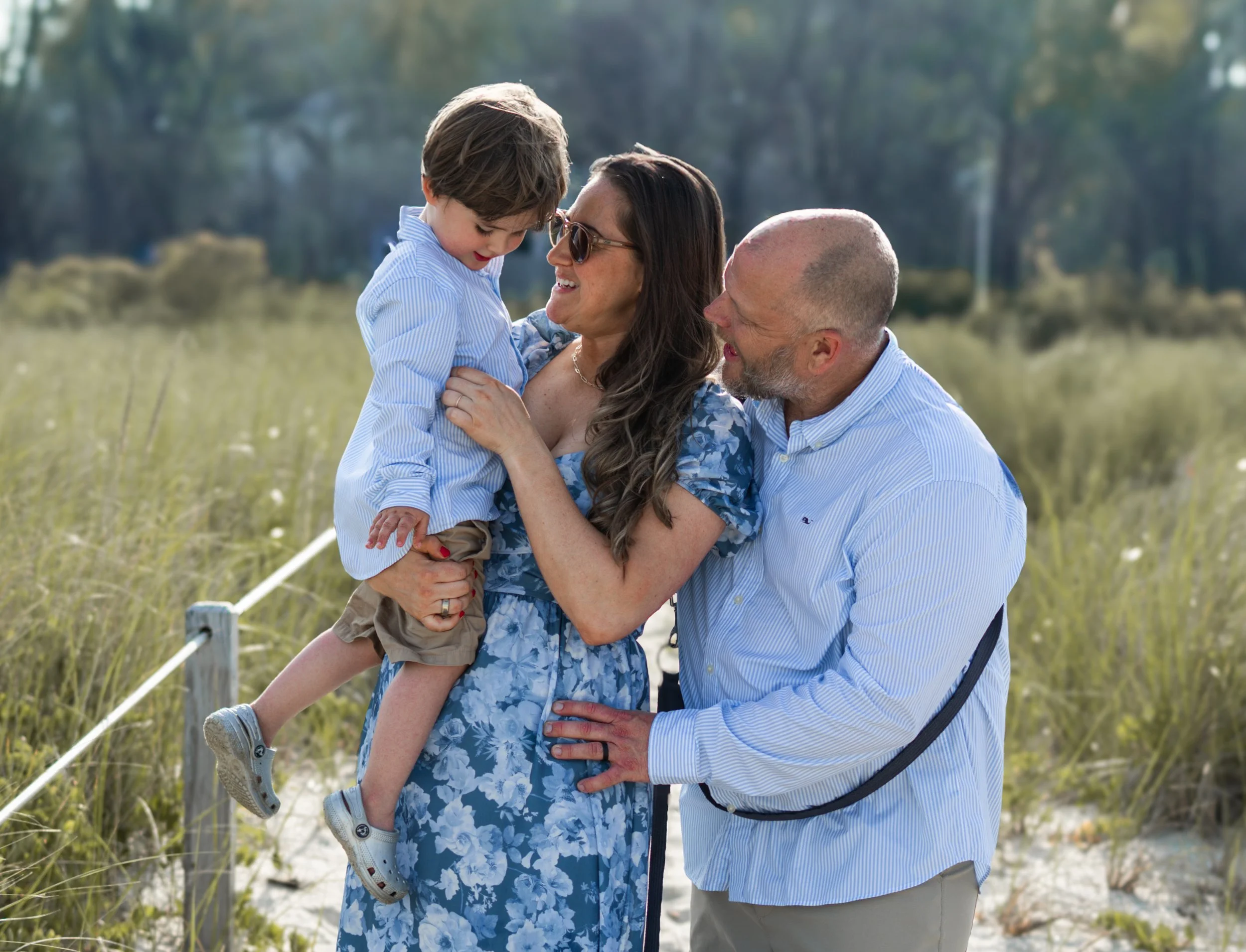 A family of three at the beach, with a woman holding a young boy in her arms, the man standing close, all smiling and enjoying each other's company.