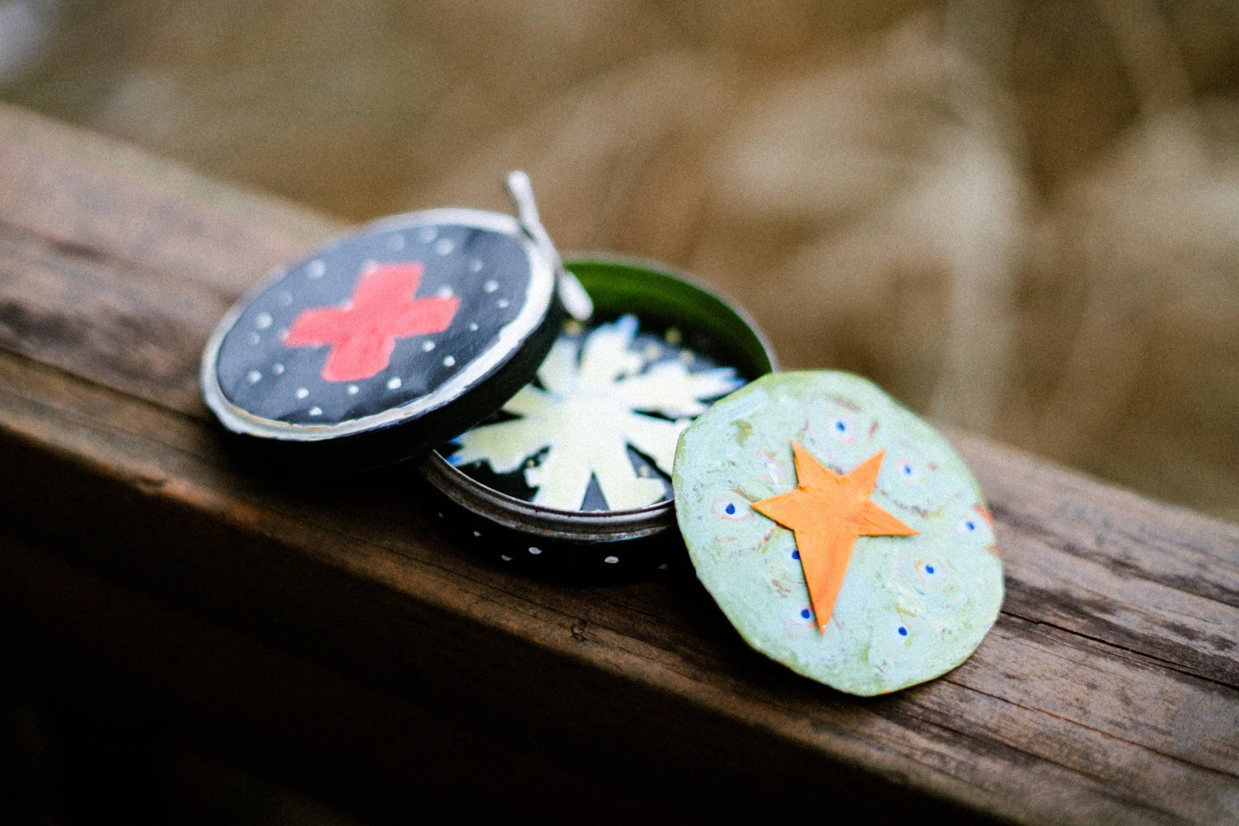 Close-up of a small round metal container with a black lid, decorated with red cross symbols, open to reveal small patches with star and snowflake designs, resting on a wooden surface.