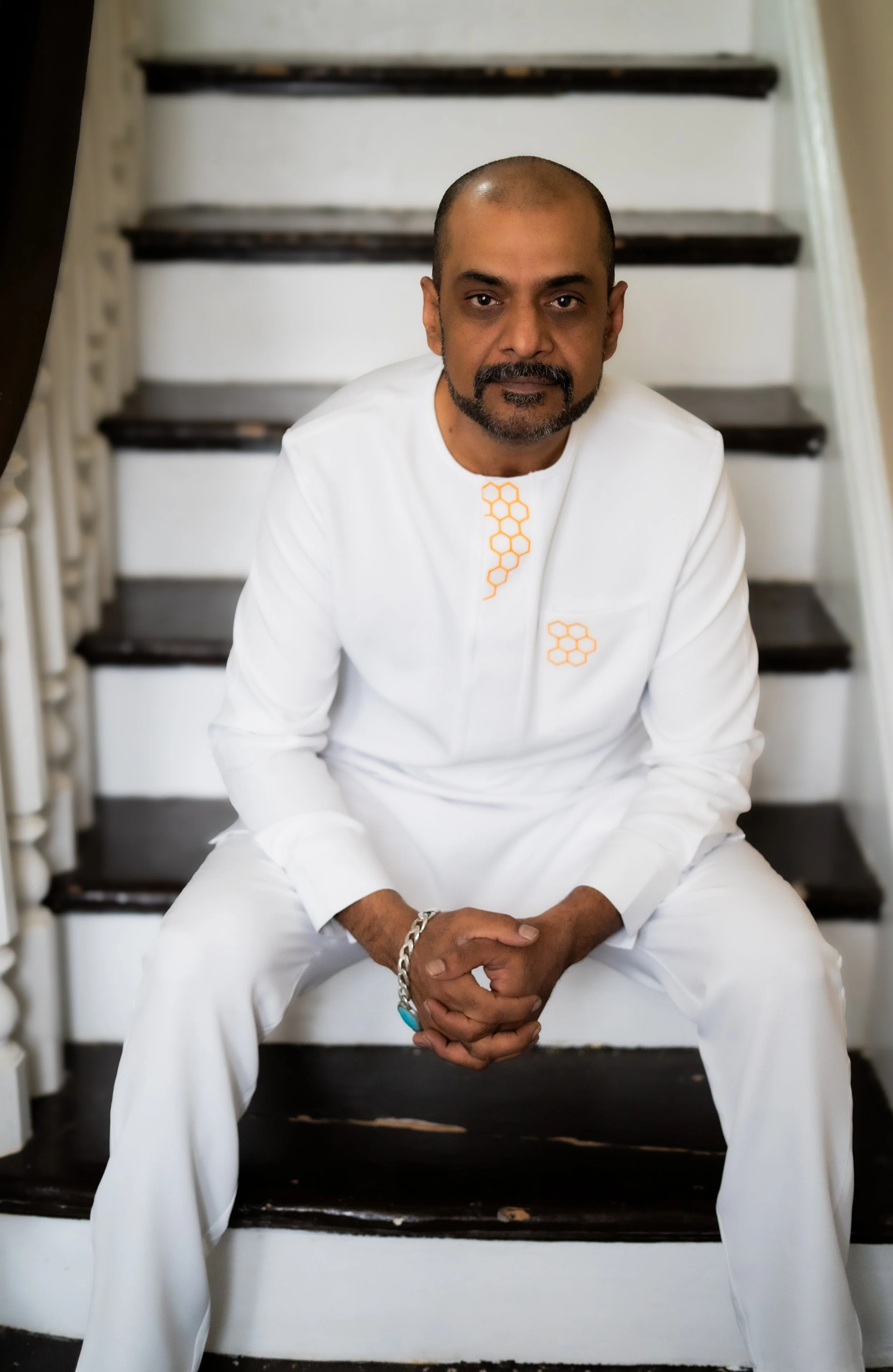 A man dressed in a white outfit with orange honeycomb patterns, sitting on a staircase with dark wooden steps and white sides, looking directly at the camera.