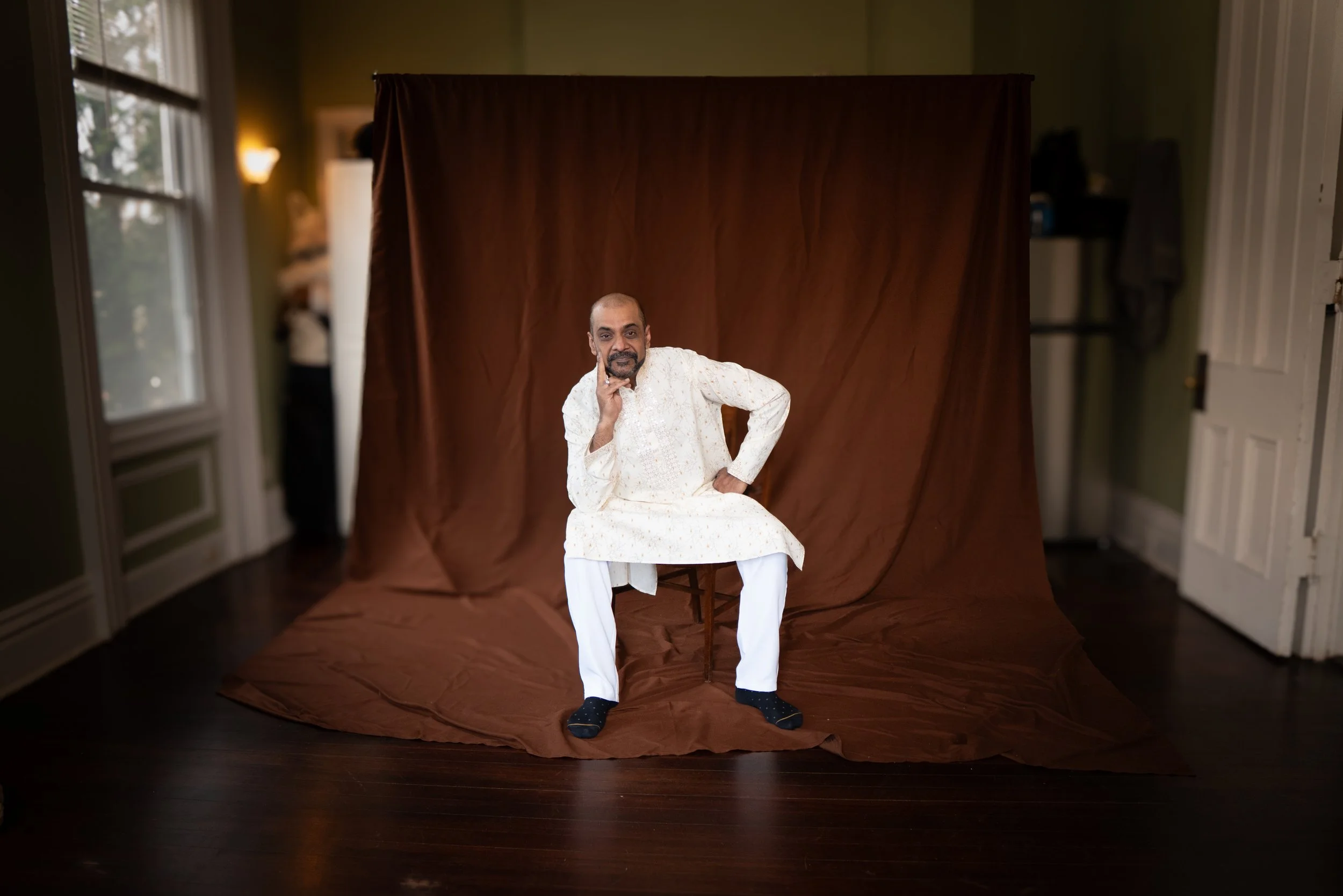 A man dressed in a traditional white Indian outfit sitting on a wooden chair in front of a brown fabric backdrop inside a room with green walls and wooden floors.