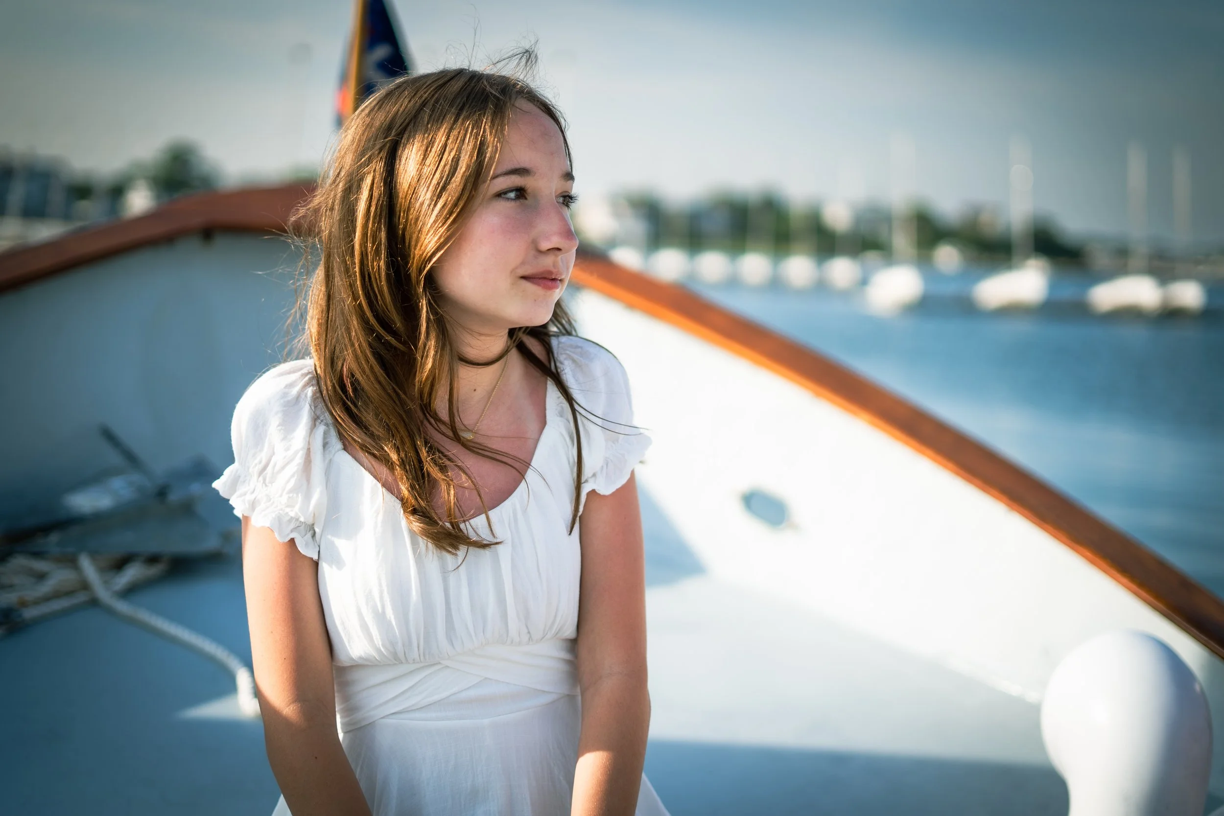 A young woman with brown hair and a white dress sitting on a boat docked in a marina, with a background of water and boats.