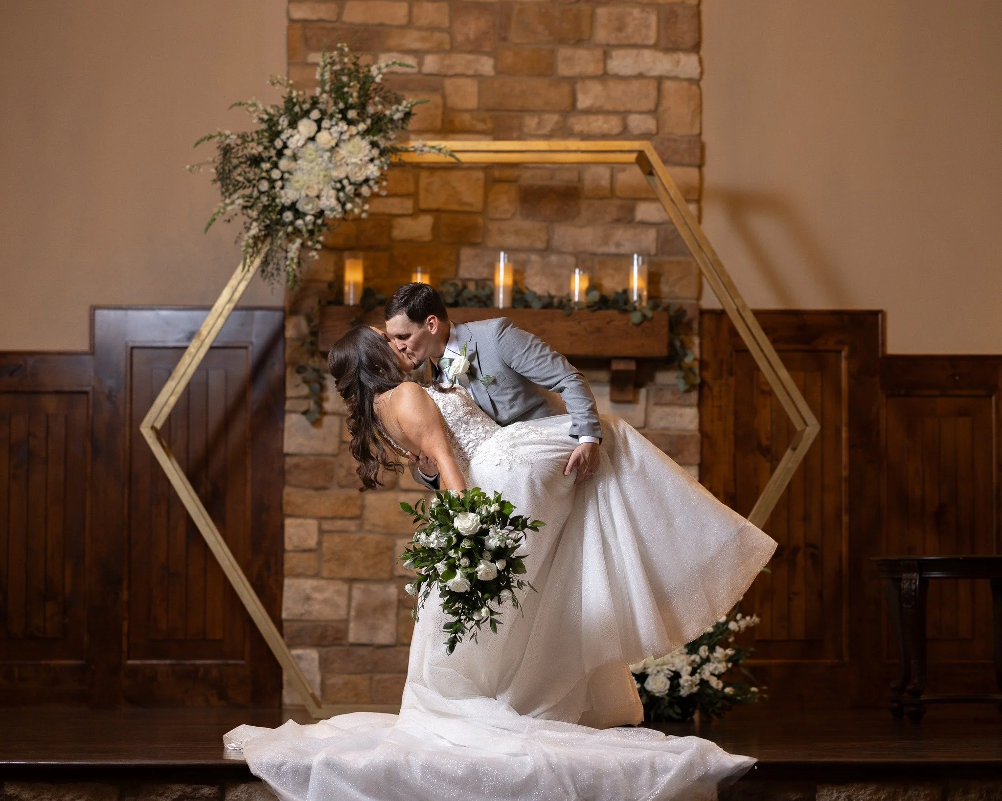 A bride and groom sharing a kiss during their wedding ceremony, with the groom dipping the bride. They are standing on a stage with a brick fireplace in the background decorated with candles and greenery, and a large floral arrangement on the left.