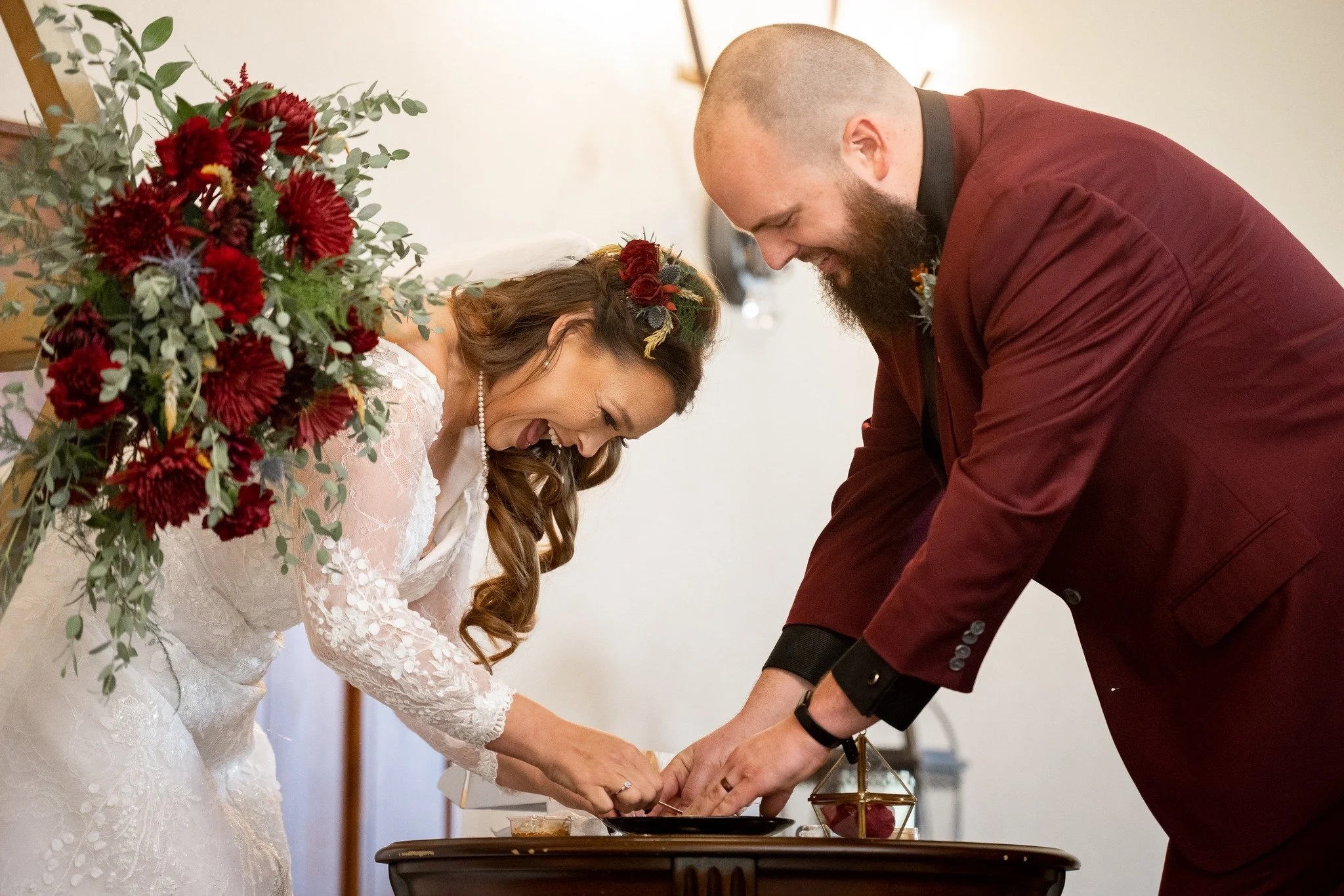 A bride and groom exchanging rings during a wedding ceremony, leaning over a table with a bouquet of red flowers nearby.