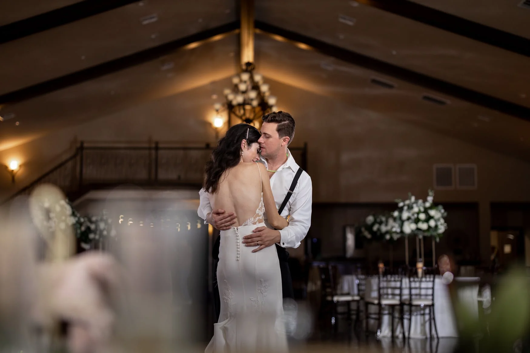 A bride and groom dancing closely at their wedding reception in an indoor venue with warm lighting, floral arrangements, and elegant decor.