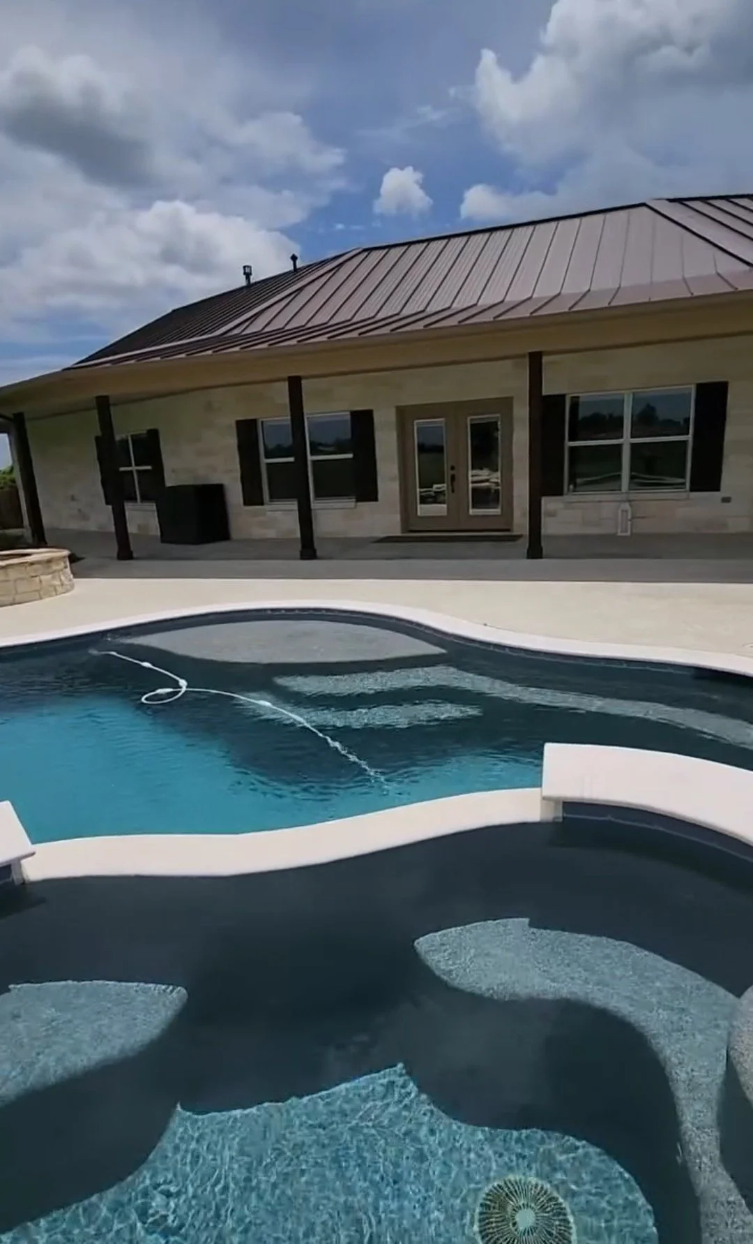 A backyard with a swimming pool in front of a house with a metal roof and beige siding, under a partly cloudy sky.