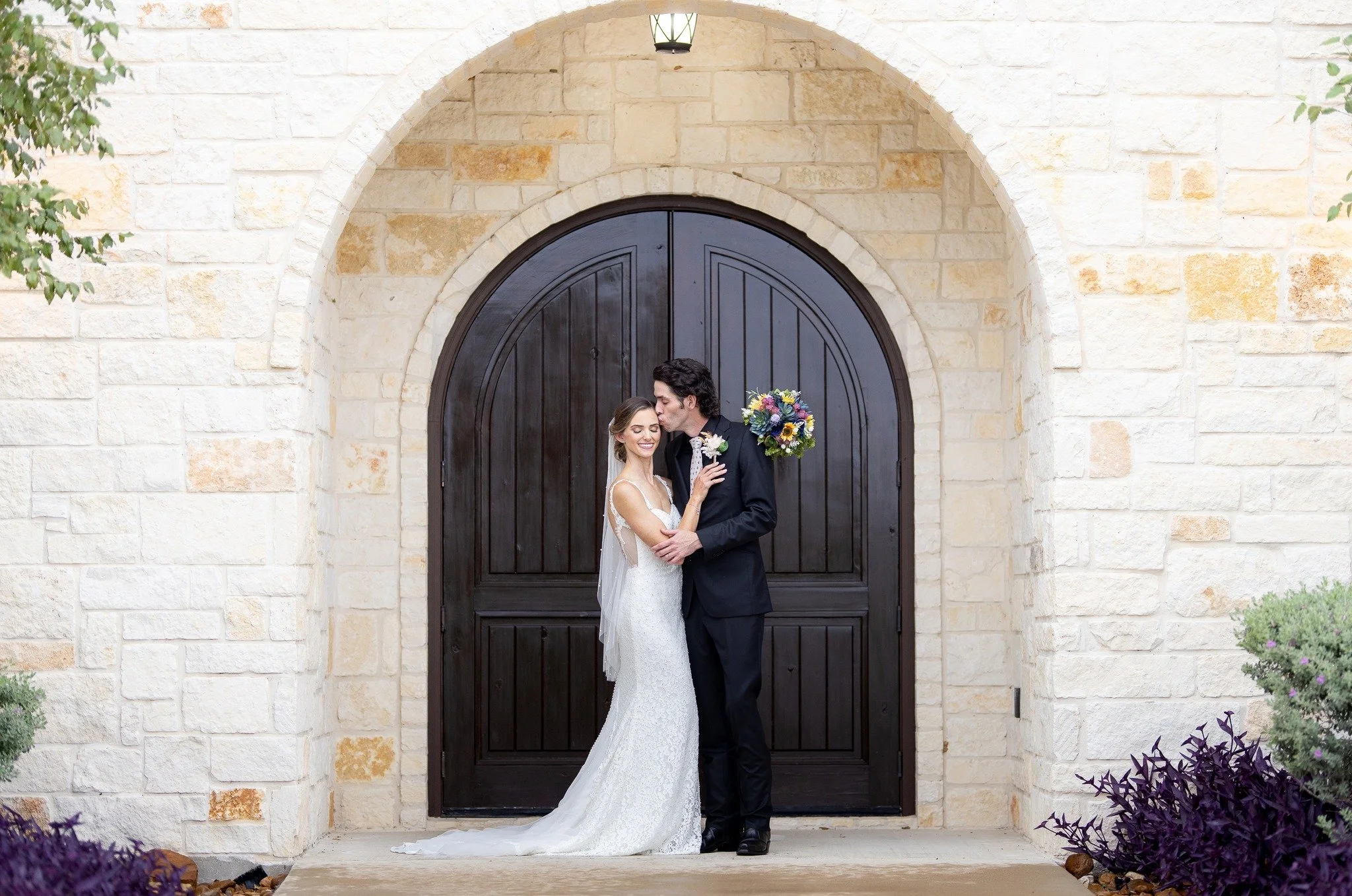 A newlywed couple stands in front of a large dark wooden arched door, embracing and smiling. The bride wears a white lace wedding dress, and the groom wears a black suit. The groom holds a colorful bouquet of flowers, and the bride has a flower bouto