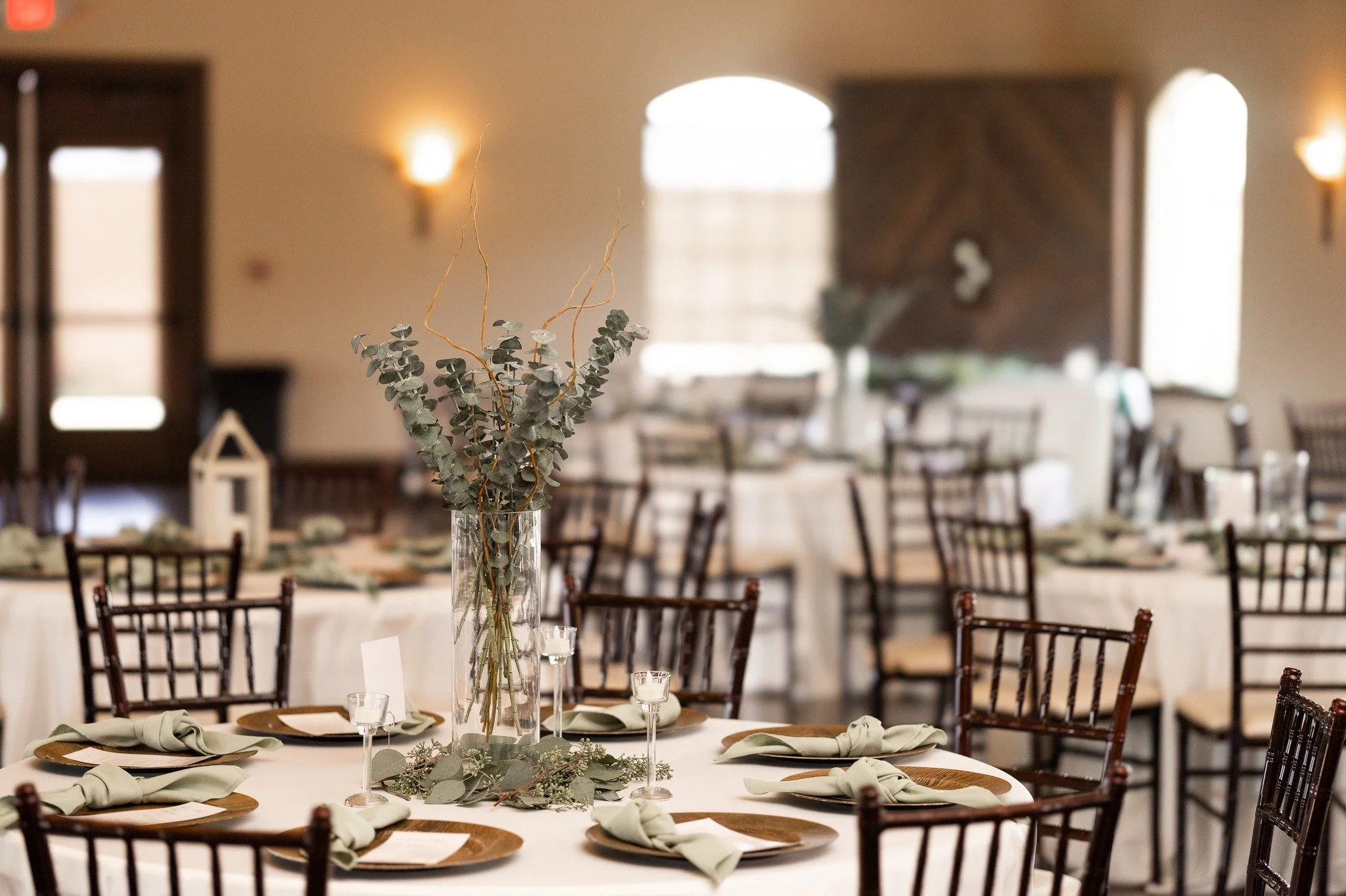 A round wedding reception table decorated with a tall glass vase filled with eucalyptus and curly willow branches, set with plates, cloth napkins, and small candles, in a warmly lit banquet hall.