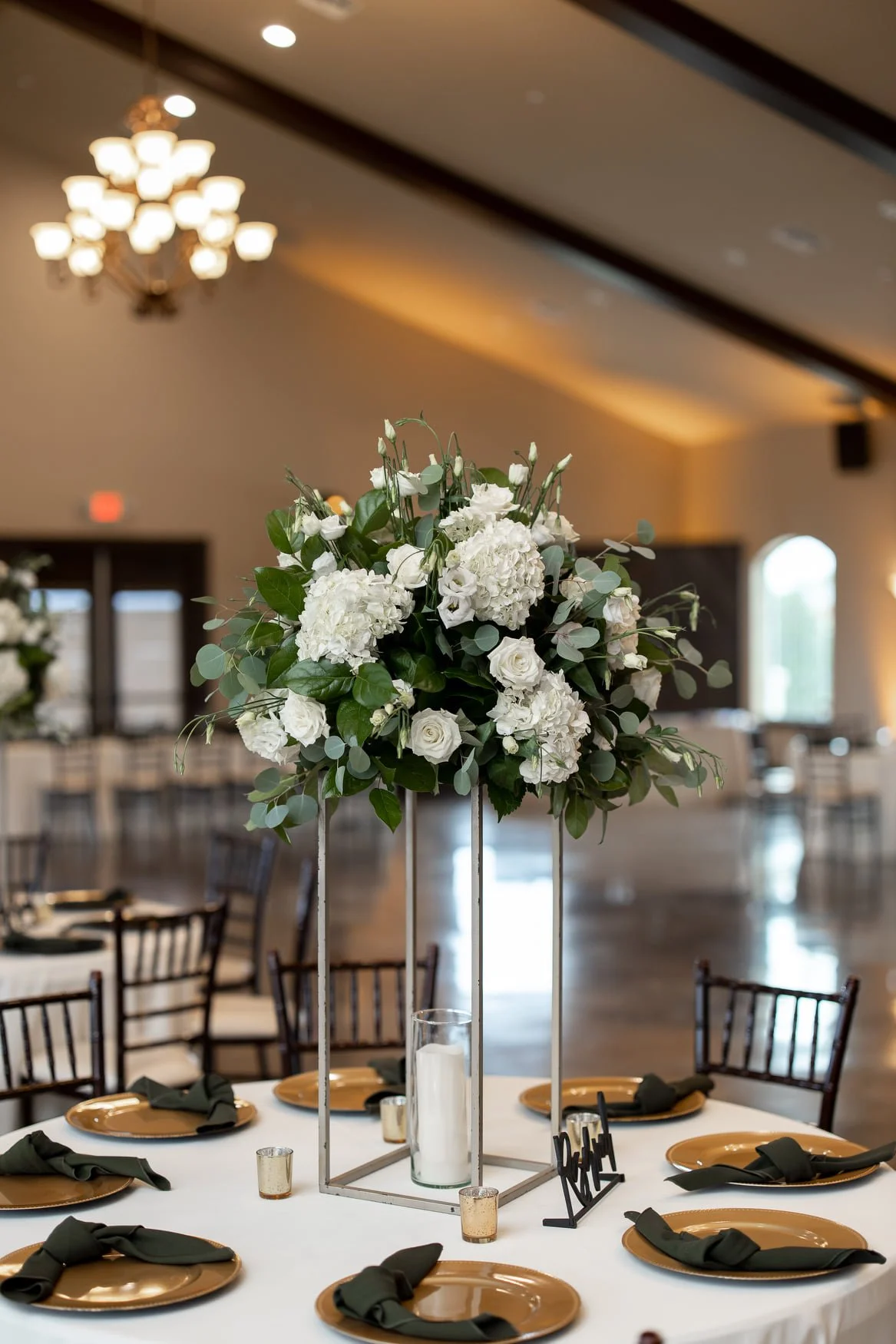Elegant banquet table centerpiece featuring a tall floral arrangement of white roses, hydrangeas, and greenery, with candles and candles holders, set in a decorated event hall.