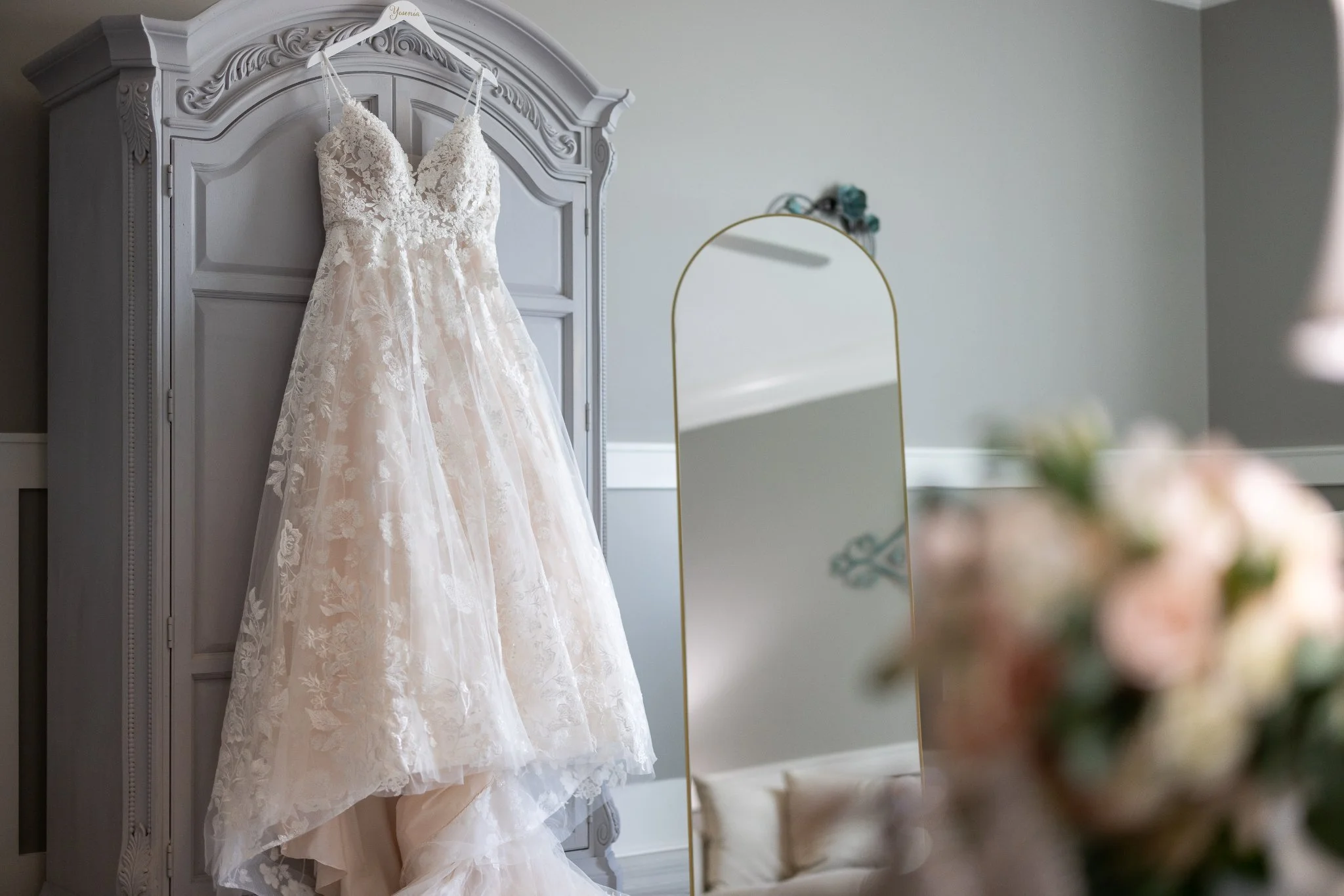 A white wedding dress hanging on an ornate gray wardrobe, reflected in a tall, gold-framed mirror, with blurred flowers in the foreground.