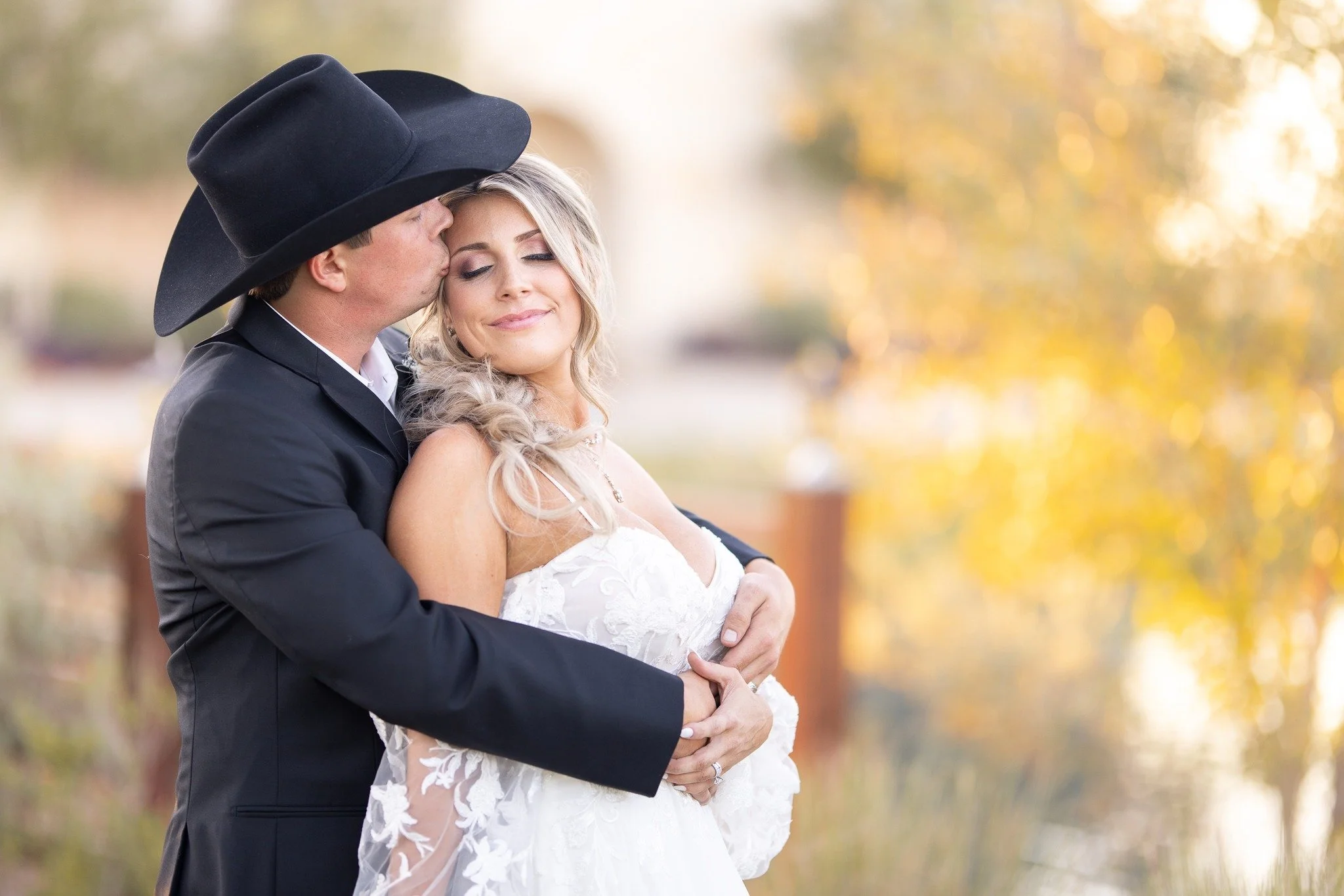 A newlywed couple embracing outdoors; the groom is kissing the bride on the forehead, and she is smiling with her eyes closed. The groom wears a black cowboy hat and tuxedo, while the bride wears a white lace wedding gown with curly blonde hair. Autu