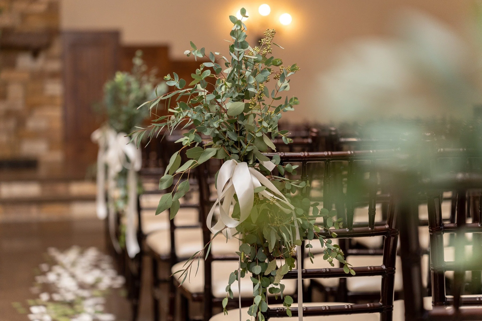 Decorative floral arrangement with greenery and white ribbons on dark wooden chairs at an interior event space.