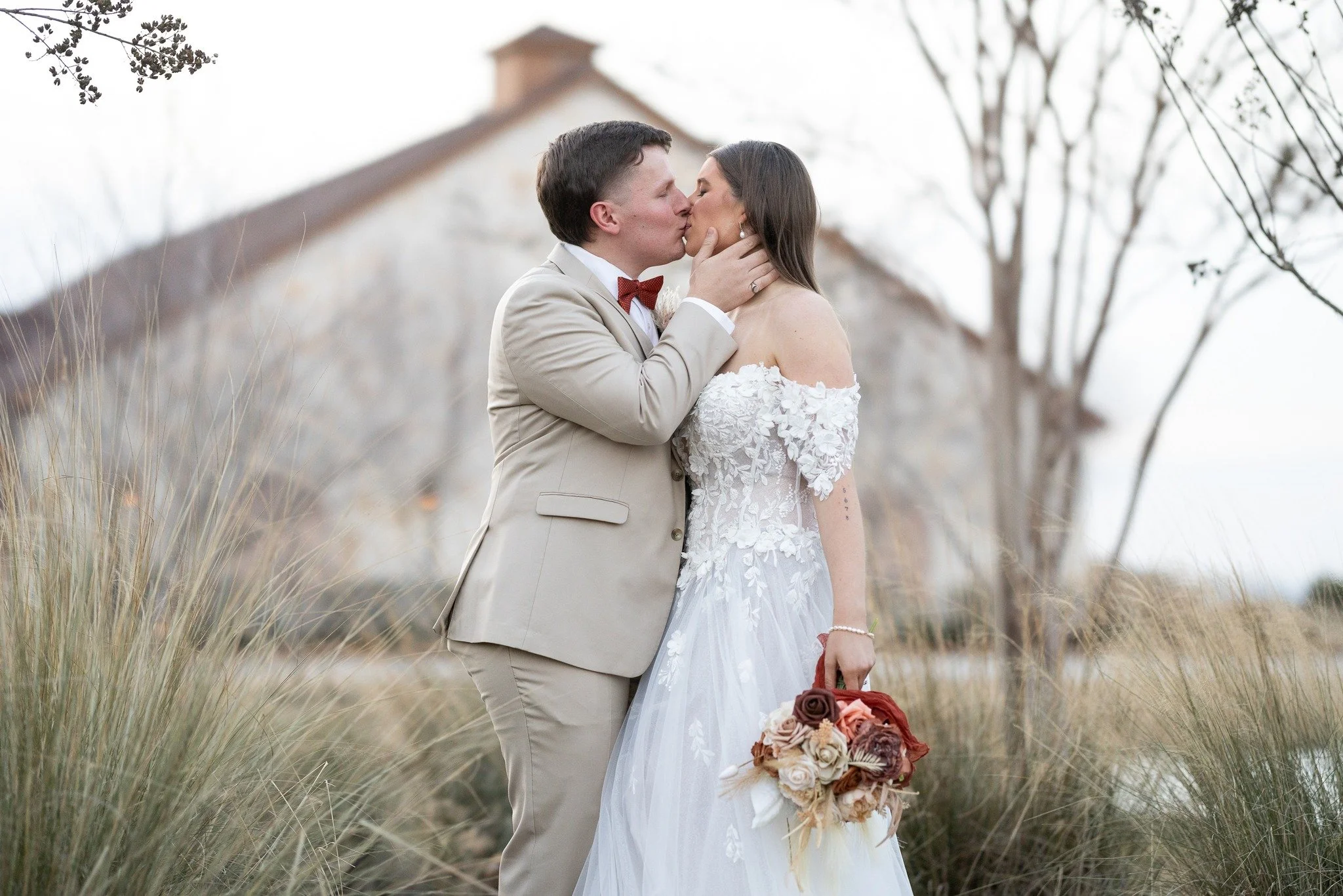 A bride and groom share a kiss outdoors, with the groom in a beige suit and red bow tie, and the bride in a white off-shoulder wedding dress holding a bouquet of flowers, with a rustic building and trees in the background.