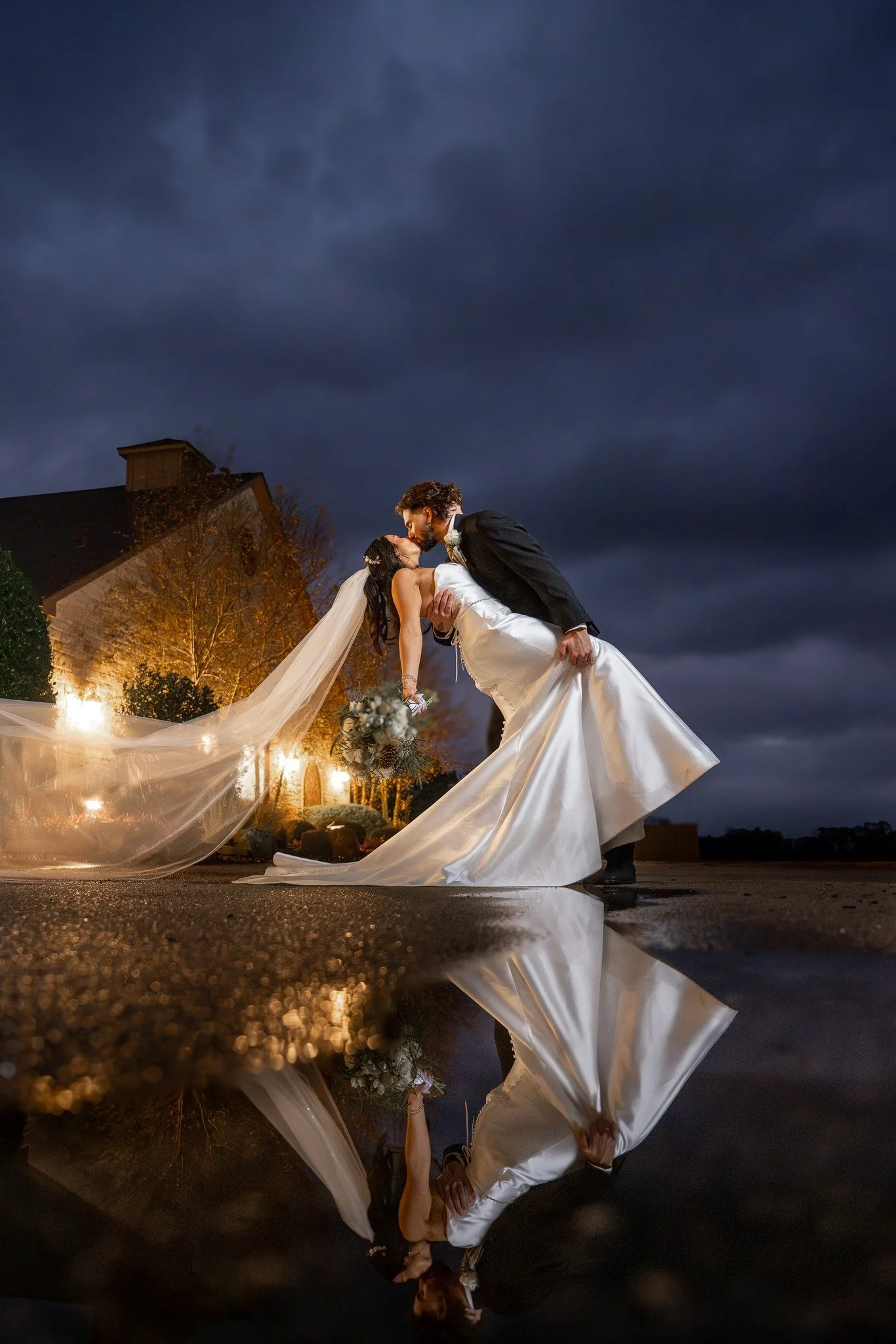A newlywed couple sharing a kiss outdoors at night, with the groom dipping the bride. The bride is holding a bouquet, and their reflection is visible in a puddle on the ground. The sky is dark with stormy clouds, and soft lighting illuminates the sce