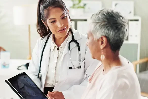 A healthcare professional, likely a doctor, is talking to an older female patient in a clinical setting, showing her information on a tablet device.