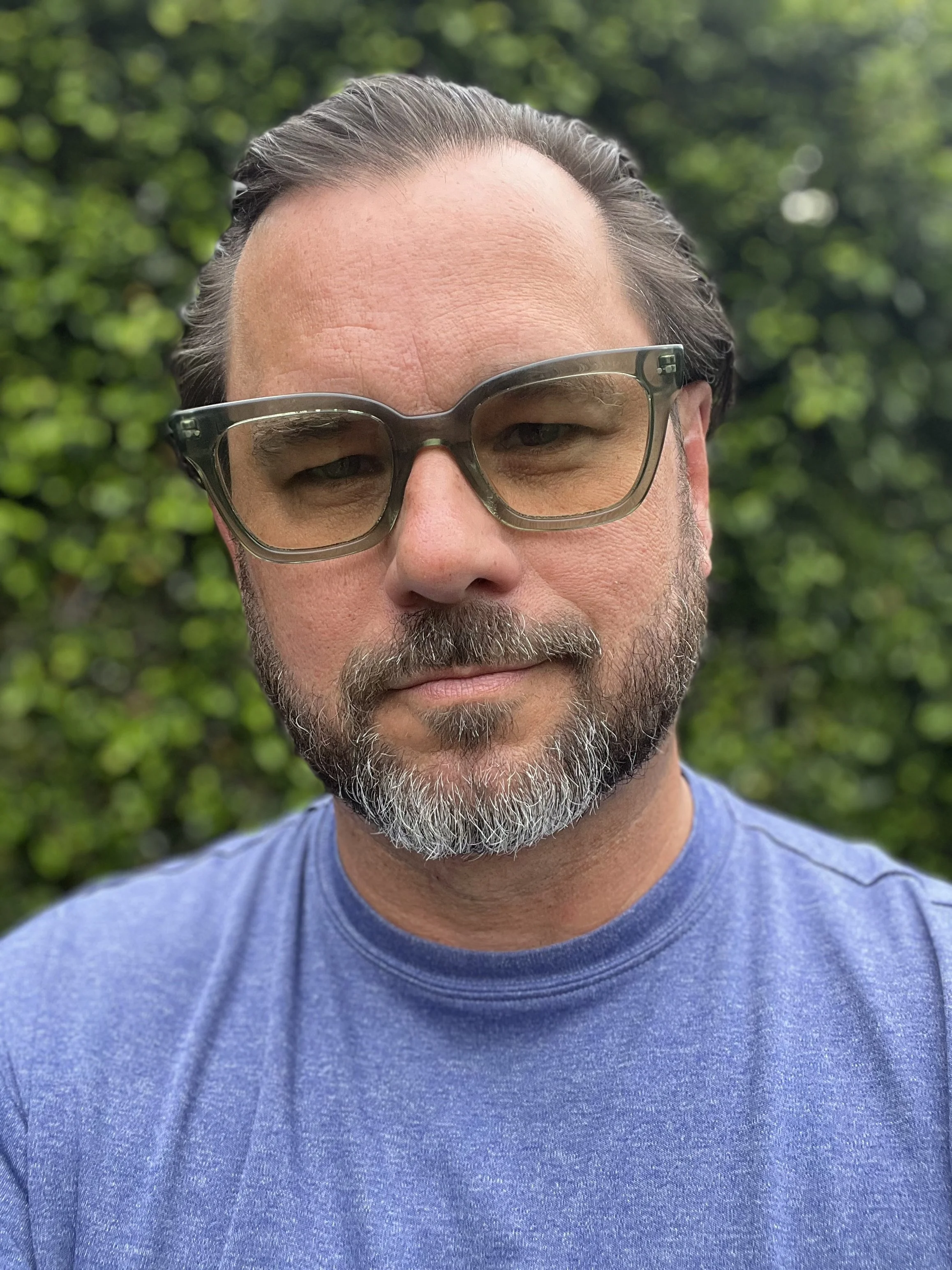 A man with dark hair, glasses, and a gray beard wearing a blue t-shirt, standing outdoors with green foliage in the background.