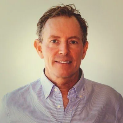 A middle-aged man with short brown hair, wearing a light purple button-down shirt, smiling at the camera, against a plain light-colored background.