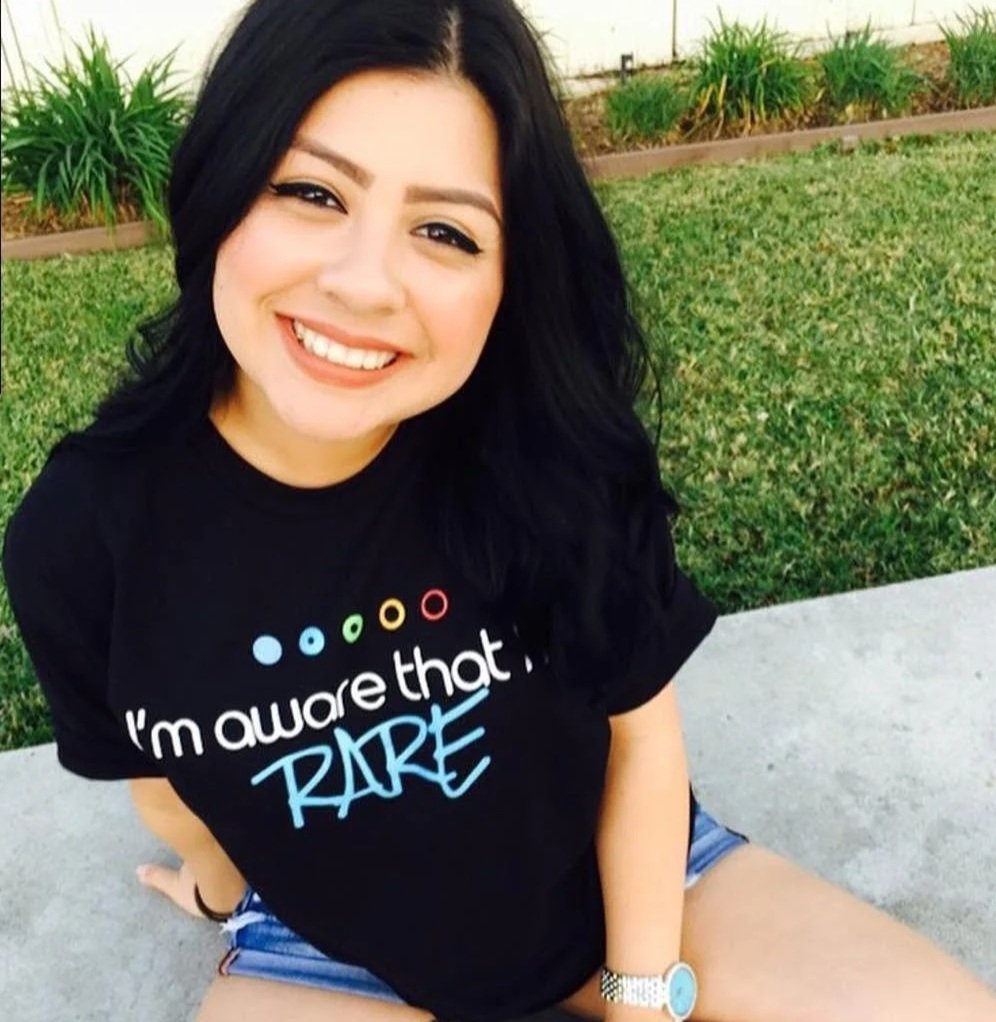 A smiling woman with black hair, wearing a black T-shirt with colorful dots and the phrase "I'm aware that I'm'" and the word "RARE" in blue, sitting on a concrete surface in front of a grassy area with plants.