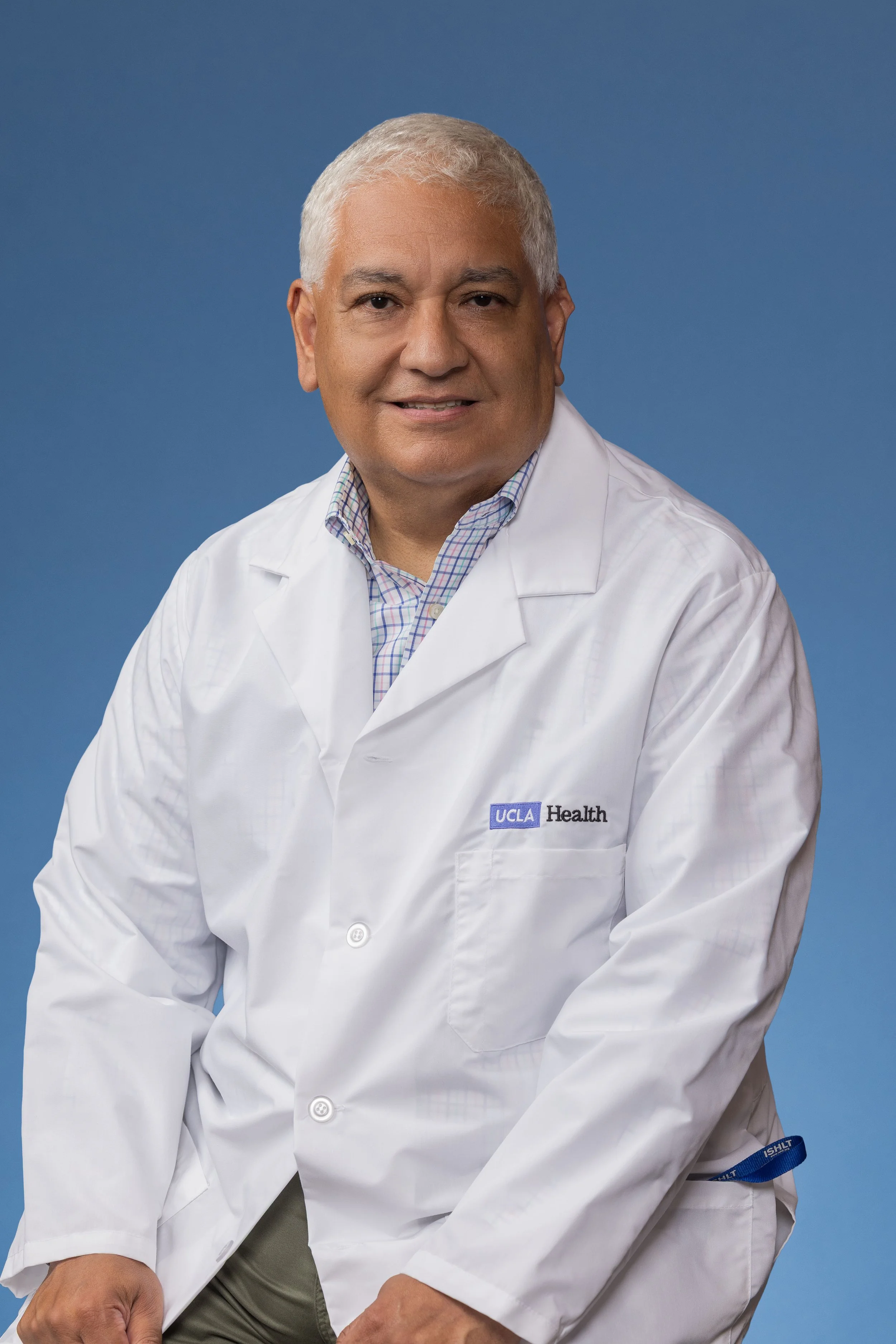 A man in a white UCLA Health lab coat sitting against a blue background.