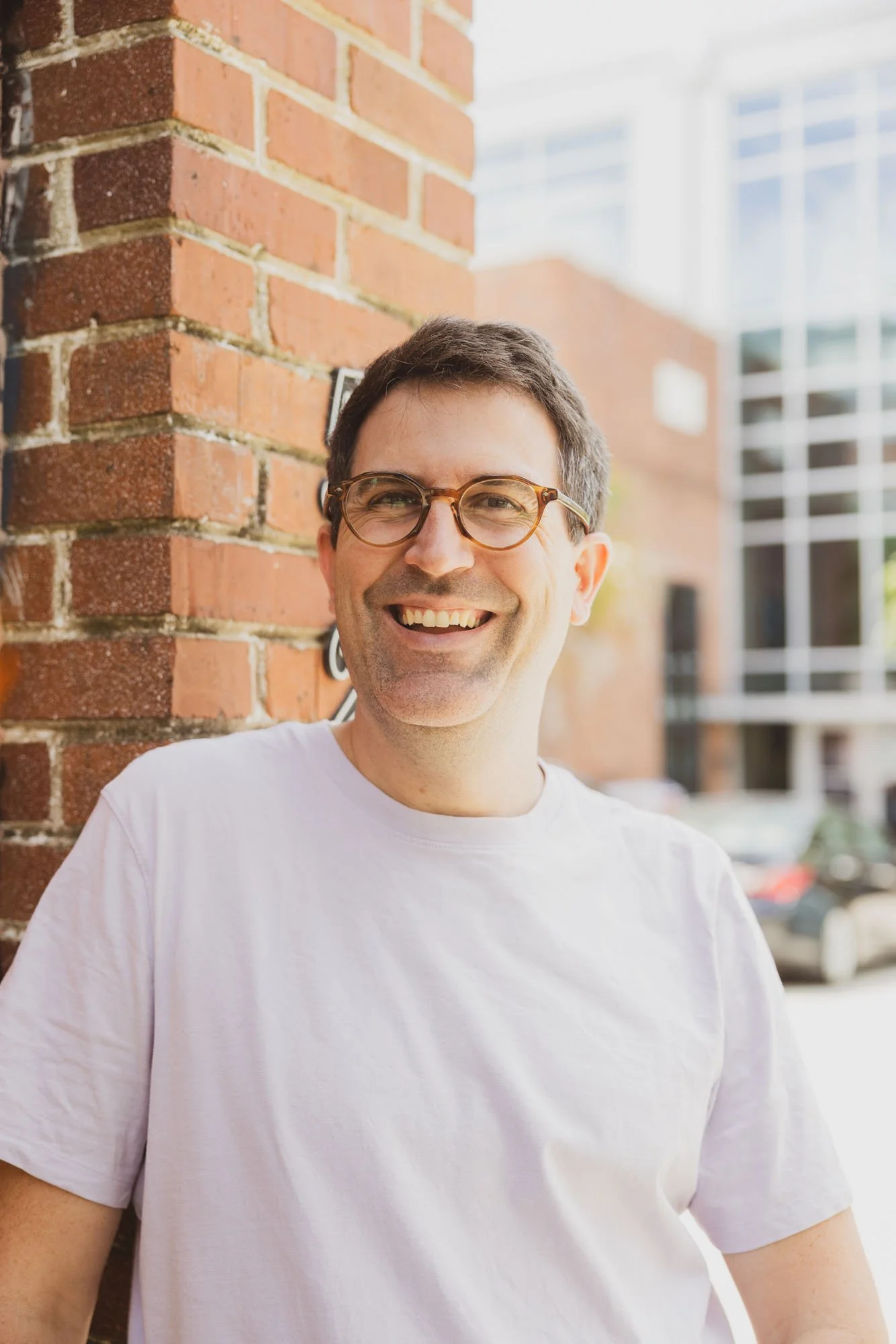 Close-up of a man with dark hair and glasses smiling, wearing a dark blue button-up shirt, against a white background.