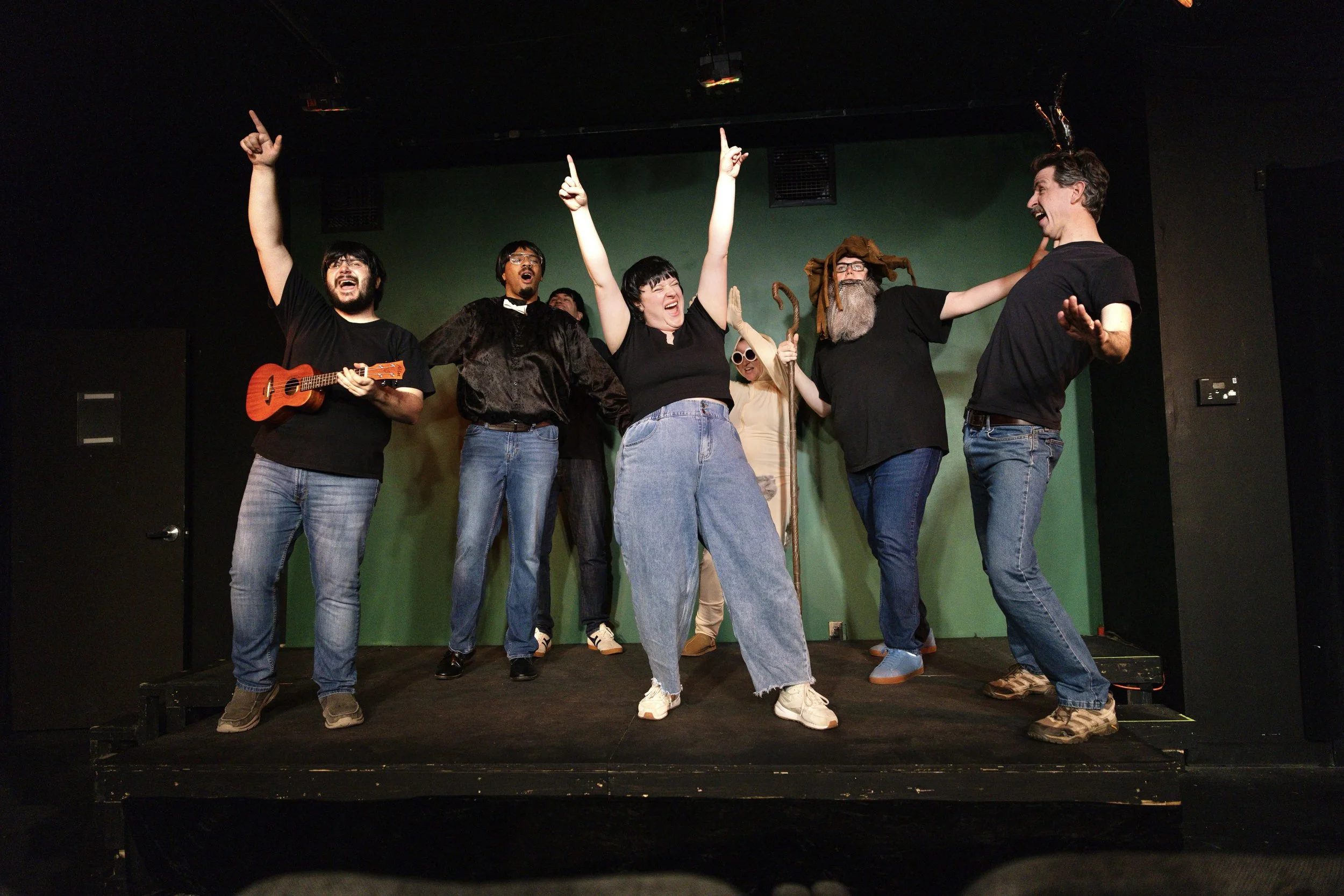 Group of six diverse people standing on a stage with a green background, celebrating with their hands raised, some holding props like a violin, glasses, and a staff with a wizard-like character, all smiling and laughing.