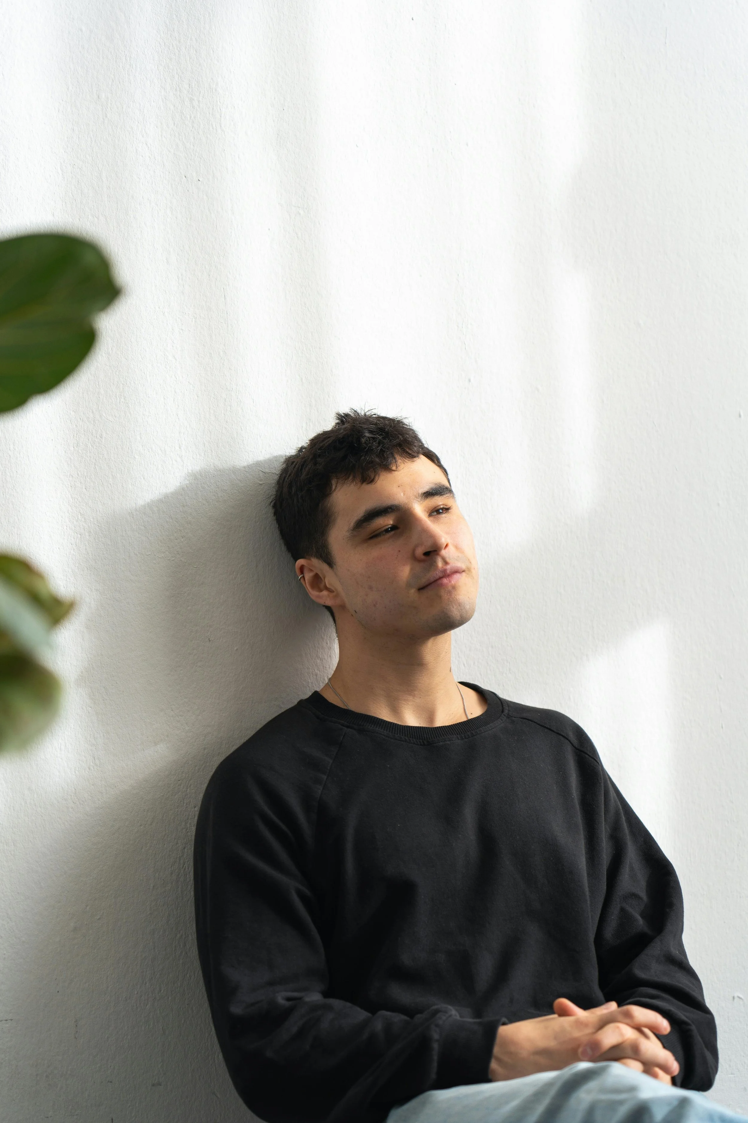 A person sitting against a white wall with a relaxed expression, wearing a black sweatshirt and light-colored pants, with a blurred green plant partially in the foreground.