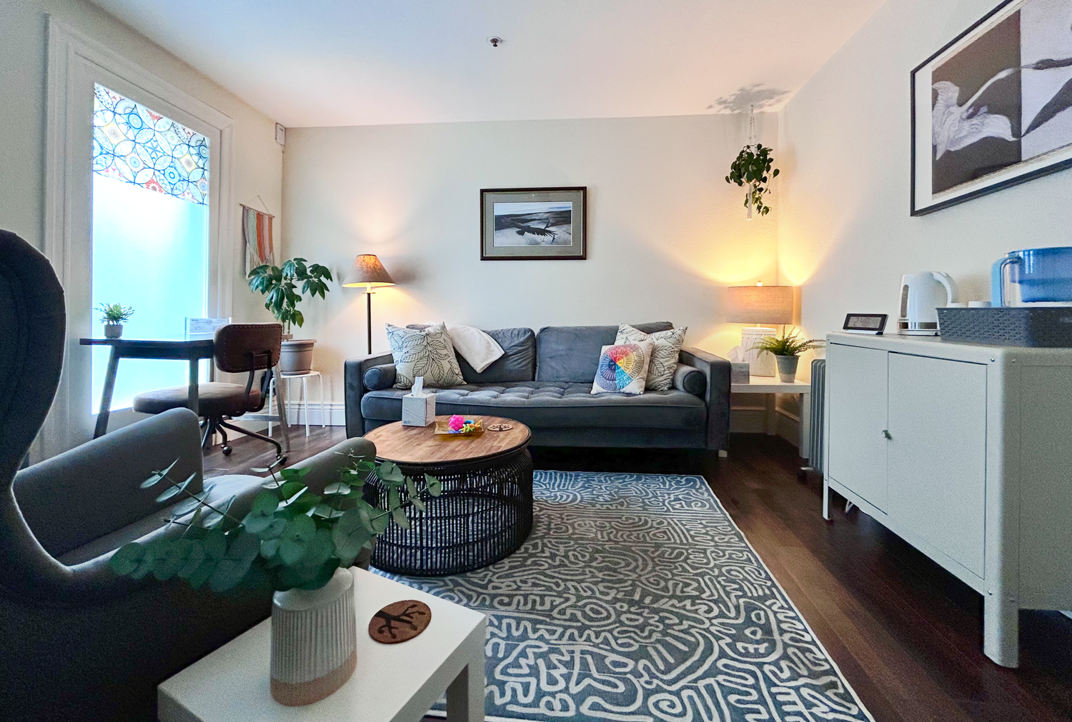 A cozy living room featuring a gray sofa with decorative pillows, a round wooden coffee table, and a patterned rug. There are two side lamps, a large air plant hanging from the ceiling, and a white cabinet with a kettle and other items. A window with a decorative curtain and a small workspace with a chair and plant are also visible.