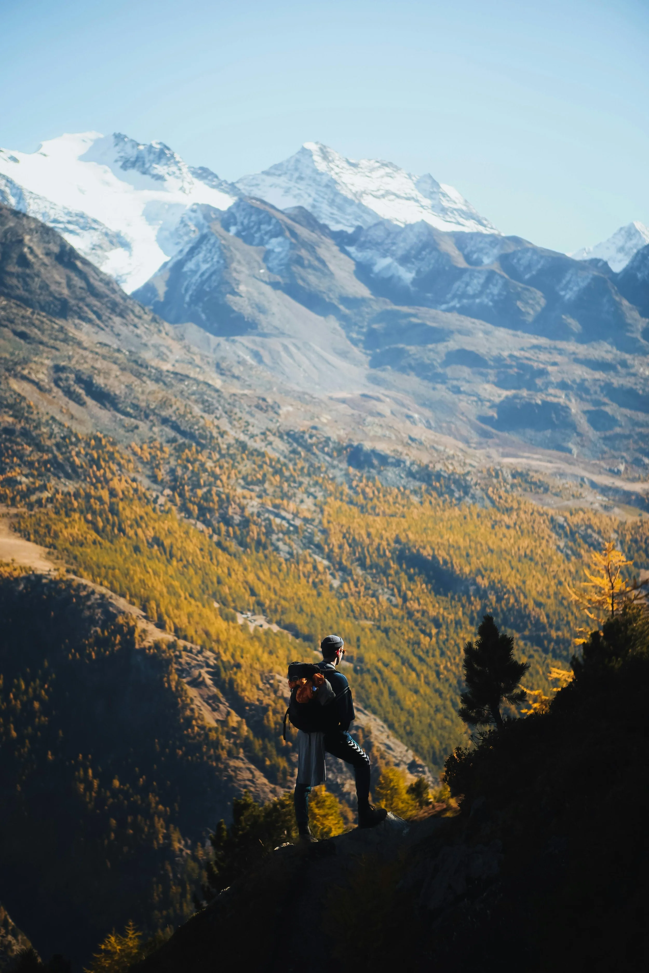 Hiker standing on a rocky ledge overlooking a mountain valley with snow-capped peaks and forested slopes during autumn.