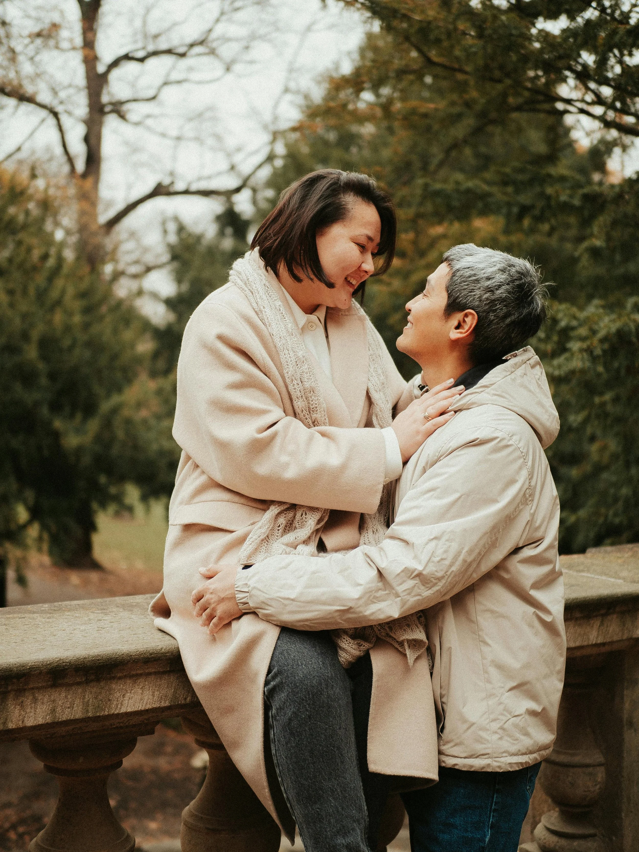 A couple enjoying a tender moment outdoors in autumn, with trees in the background. The woman is sitting on a stone bench, smiling, while a woman kneels in front of her, holding her hands and looking up at her.
