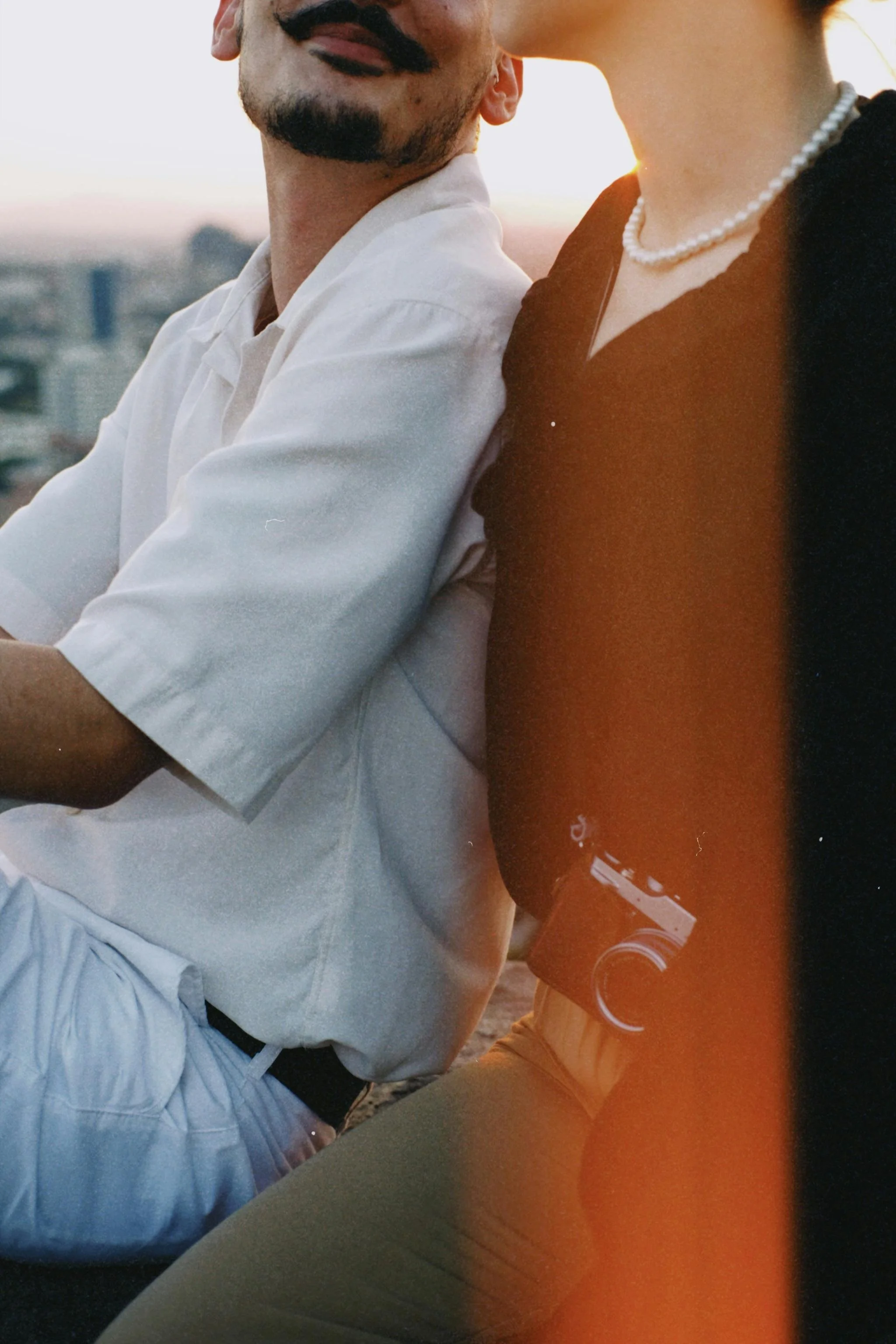 A close-up of couple sitting outdoors with a cityscape in the background during sunset. One person is wearing a white shirt, and the other is wearing a black top, a pearl necklace, and holding a camera.