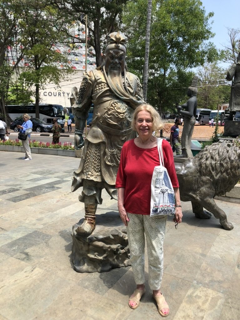 A woman in a red top and patterned pants standing next to a large warrior statue. The statue is ornately decorated and set in an outdoor area with trees and a building in the background.