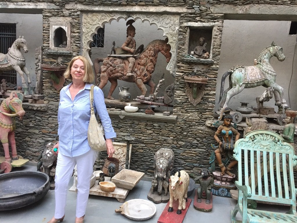 A woman stands in front of a display of carved wooden statues and sculptures, including animals and figures, with a stone wall backdrop. She is smiling and holding a tote bag, wearing a light blue shirt and white pants.