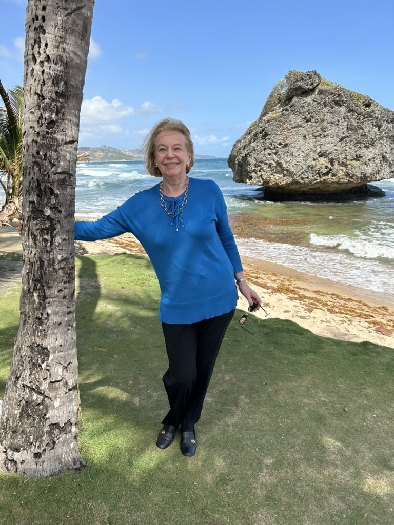 Woman in blue sweater standing by palm tree on a beach with large rock and ocean waves in background.