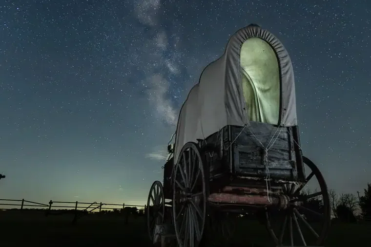 An old covered wagon with a white canvas top under a night sky filled with stars and the Milky Way.