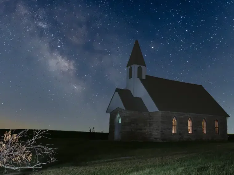 A small white church with a steeple at night, illuminated from inside, against a starry sky with the Milky Way galaxy visible.