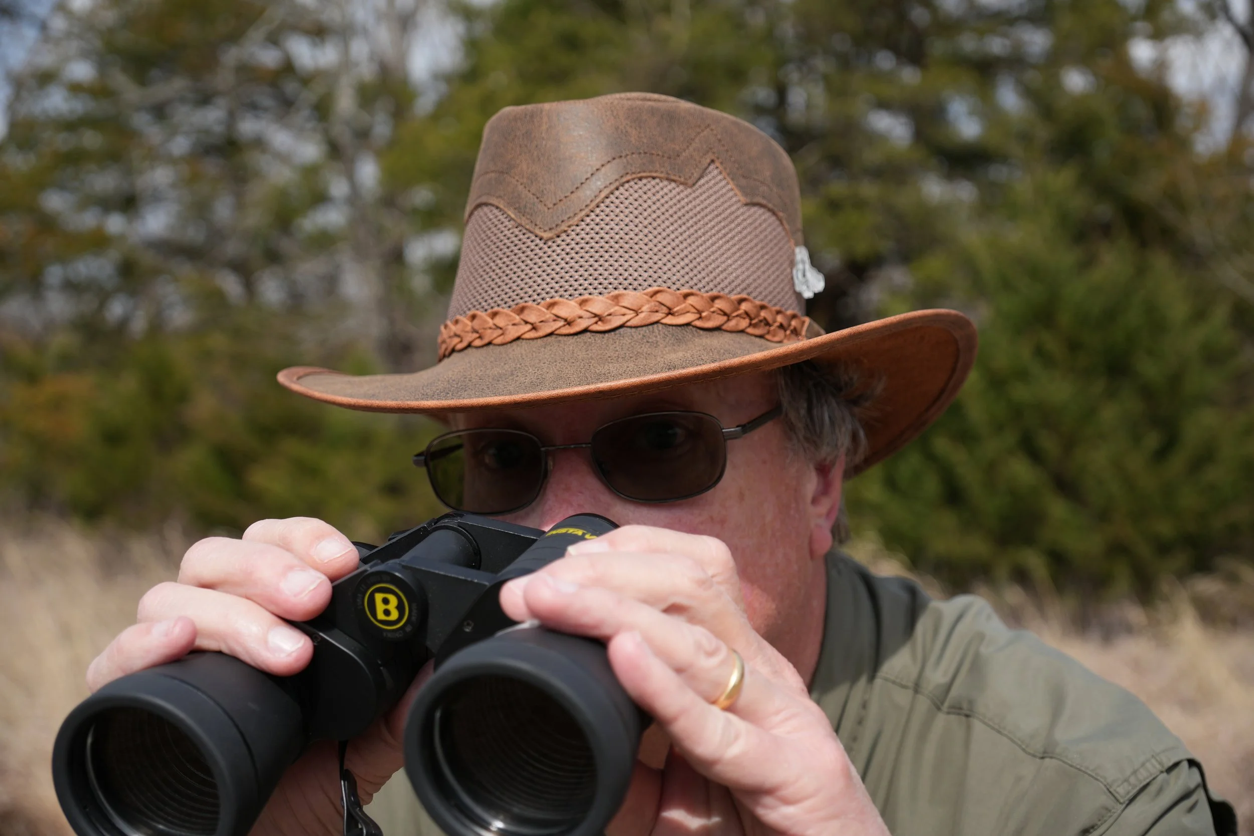 A person wearing a cowboy hat and sunglasses looking through binoculars outdoors.