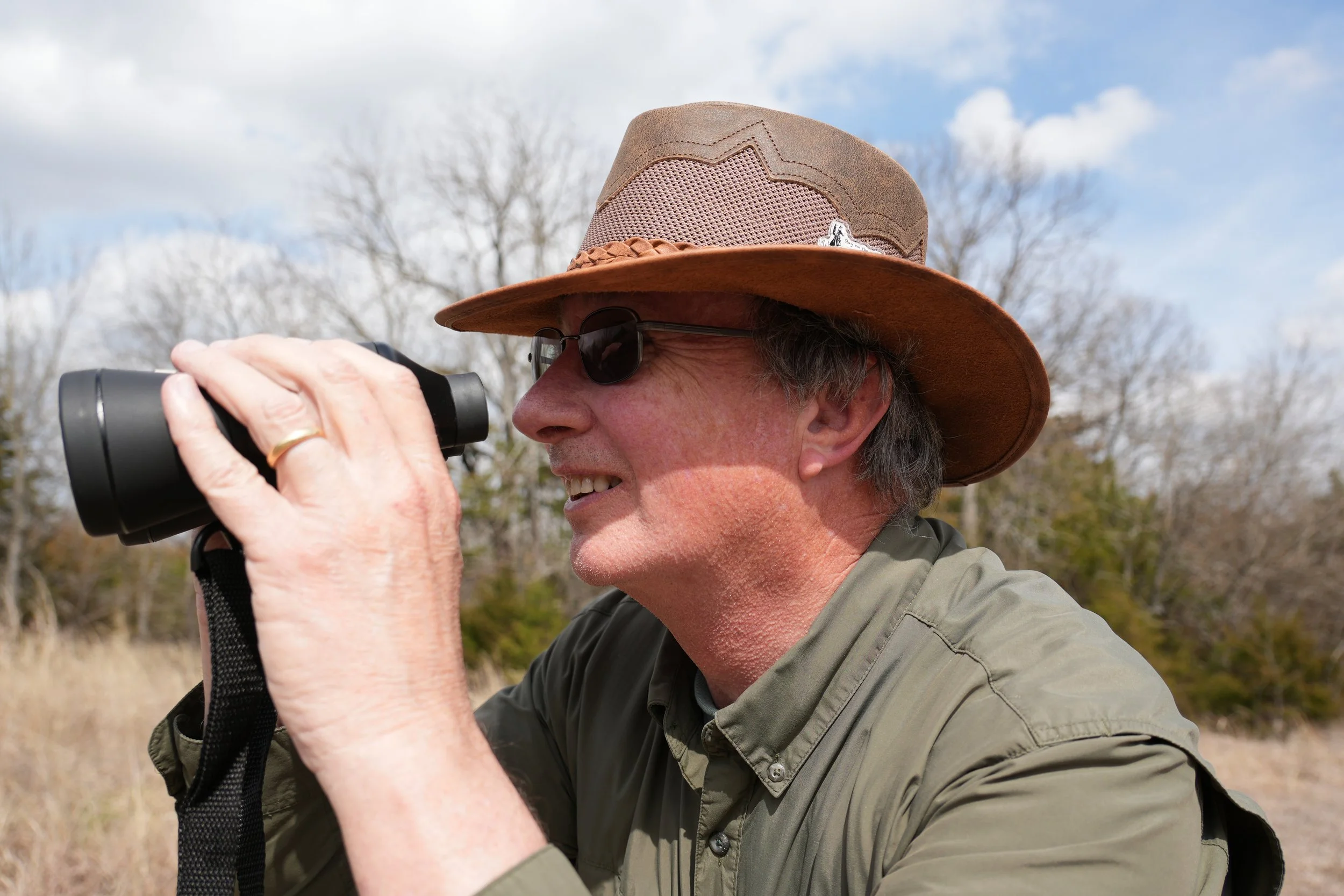 A man wearing a wide-brimmed hat, sunglasses, and a green jacket, holding binoculars up to his eyes outdoors during daytime with bare trees and a partly cloudy sky in the background.