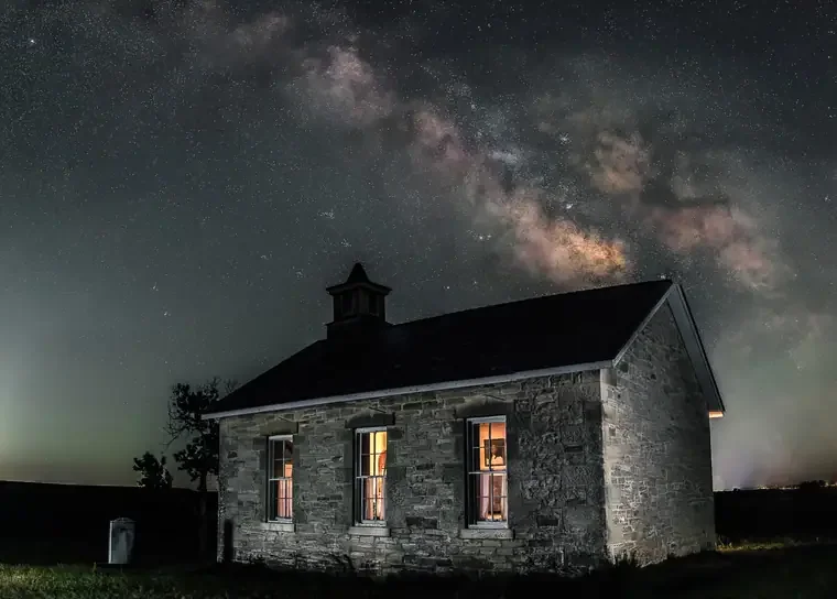 Stone house at night with three lit windows and a visible roof, under a starry sky featuring the Milky Way galaxy.