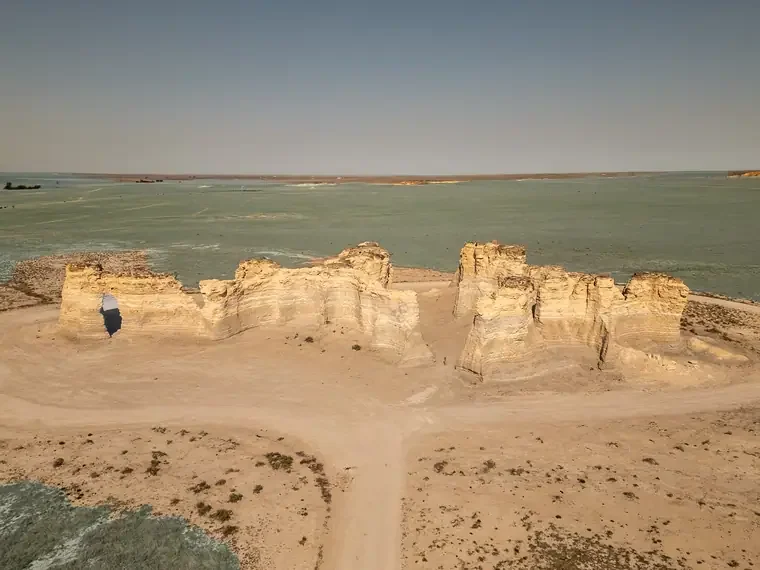 A natural rock formation with a hole in one of the rocks, set in a desert landscape near a body of water.
