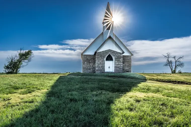 A small church with stone walls and white trim on a grassy hill, with the sun shining behind its steeple against a blue sky with clouds.