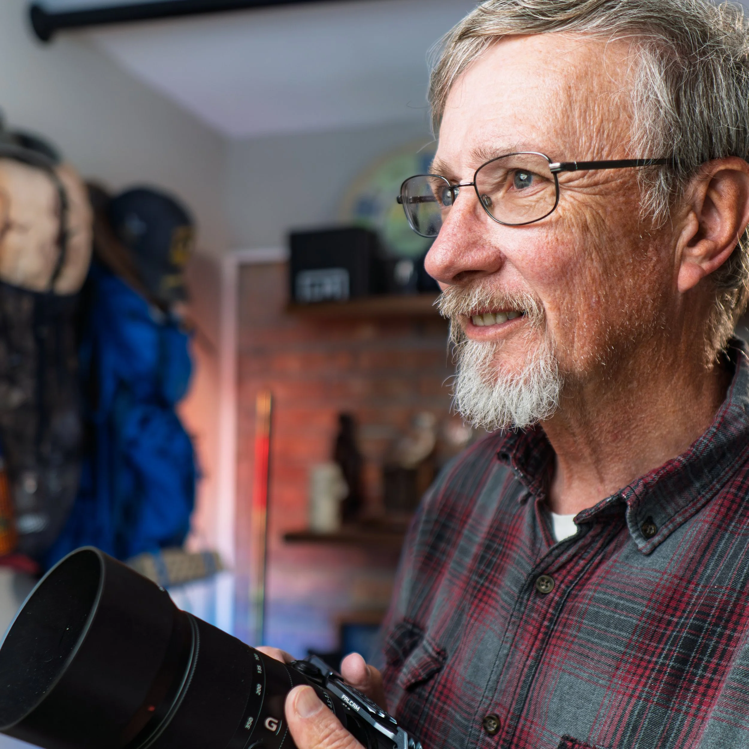 Close-up of an elderly man with gray hair, a beard, and glasses smiling while looking through a camera lens held in his hand, wearing a plaid shirt.
