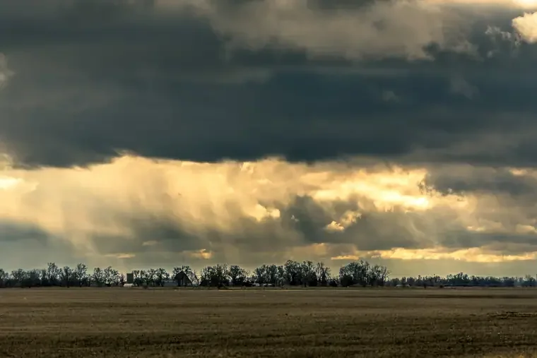A vast open field with dry grass, a line of trees in the distance, under a dramatic cloudy sky with rays of sunlight breaking through.