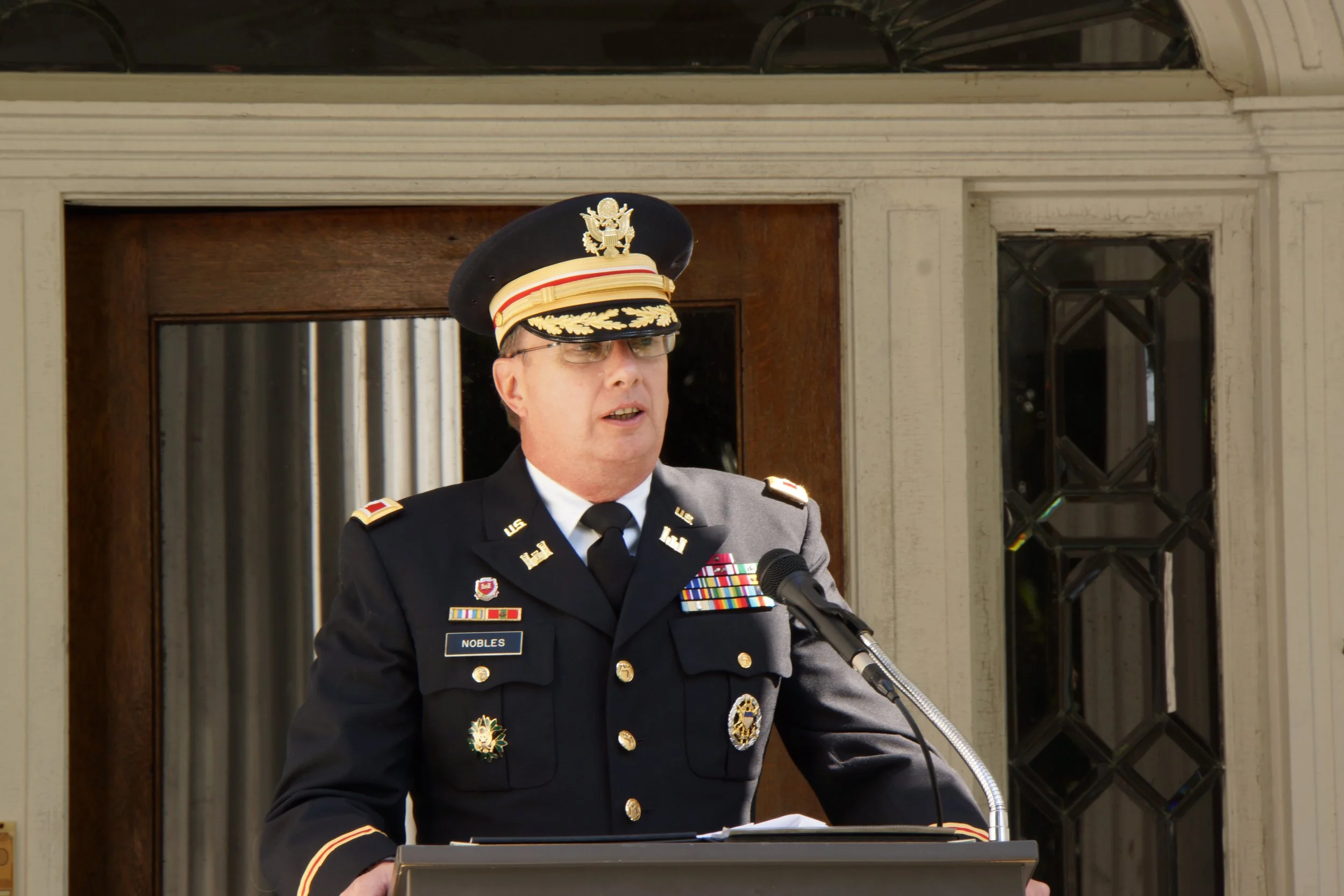 A United States Army officer in formal dress uniform speaking at a podium outdoors.