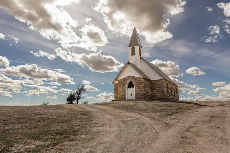 A small white and stone church with a tall steeple on a hill under a partly cloudy sky.