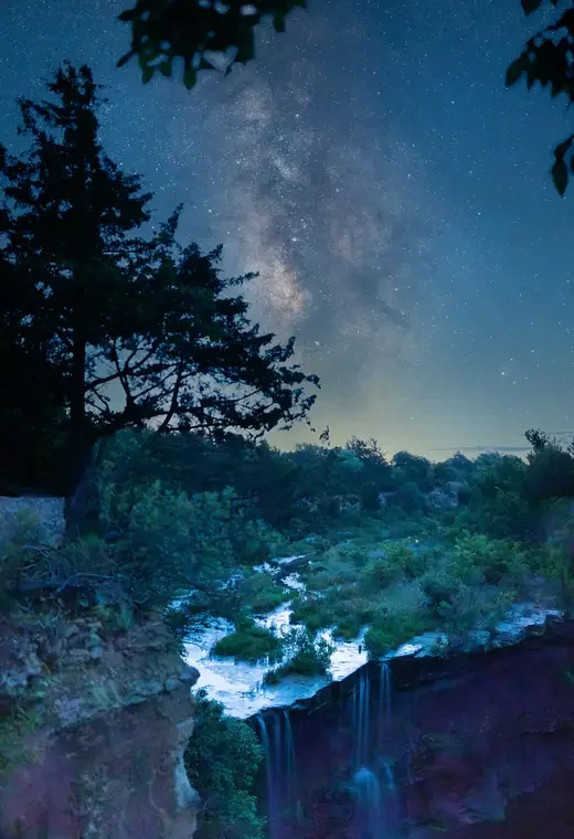 Night sky with stars and the Milky Way over a forested landscape with a small waterfall and flowing stream.