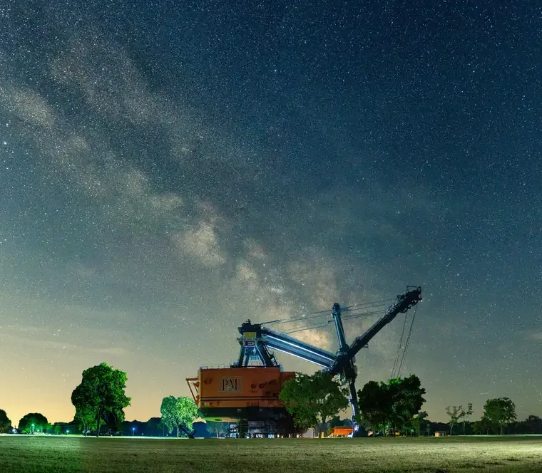 Night sky with visible stars and the Milky Way galaxy above a field with a large construction crane and trees.