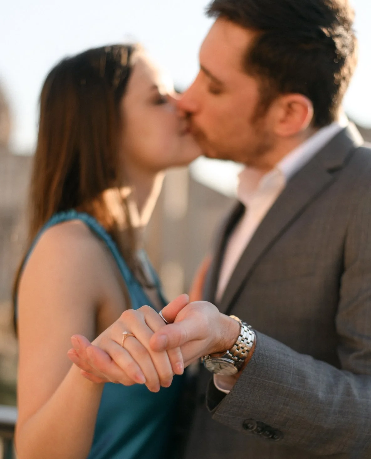 A couple kissing while holding hands, with a woman wearing a teal dress and a man in a gray suit, outdoors during sunset.