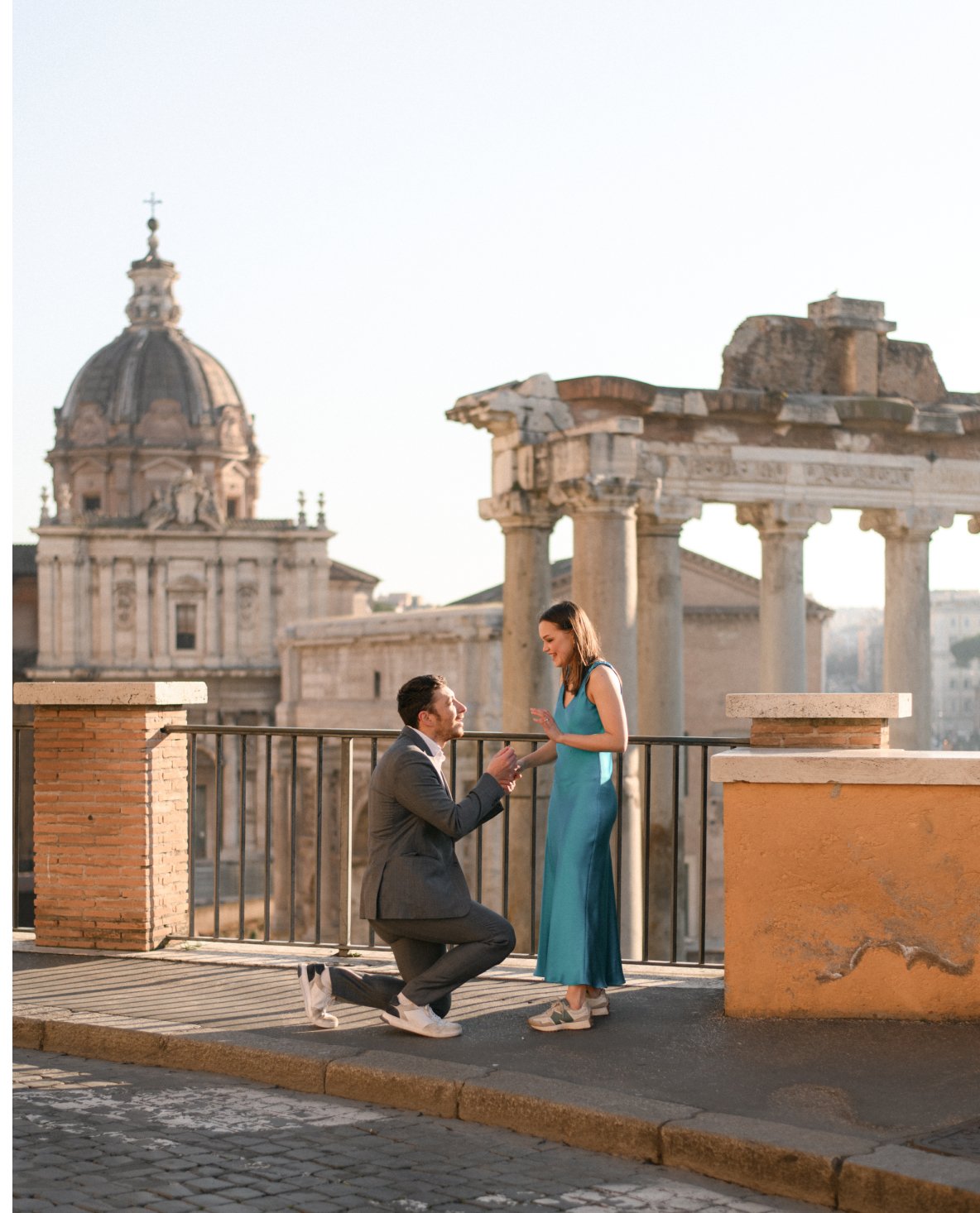 A man proposing marriage to a woman on a rooftop with ancient ruins and a dome in the background during sunset.